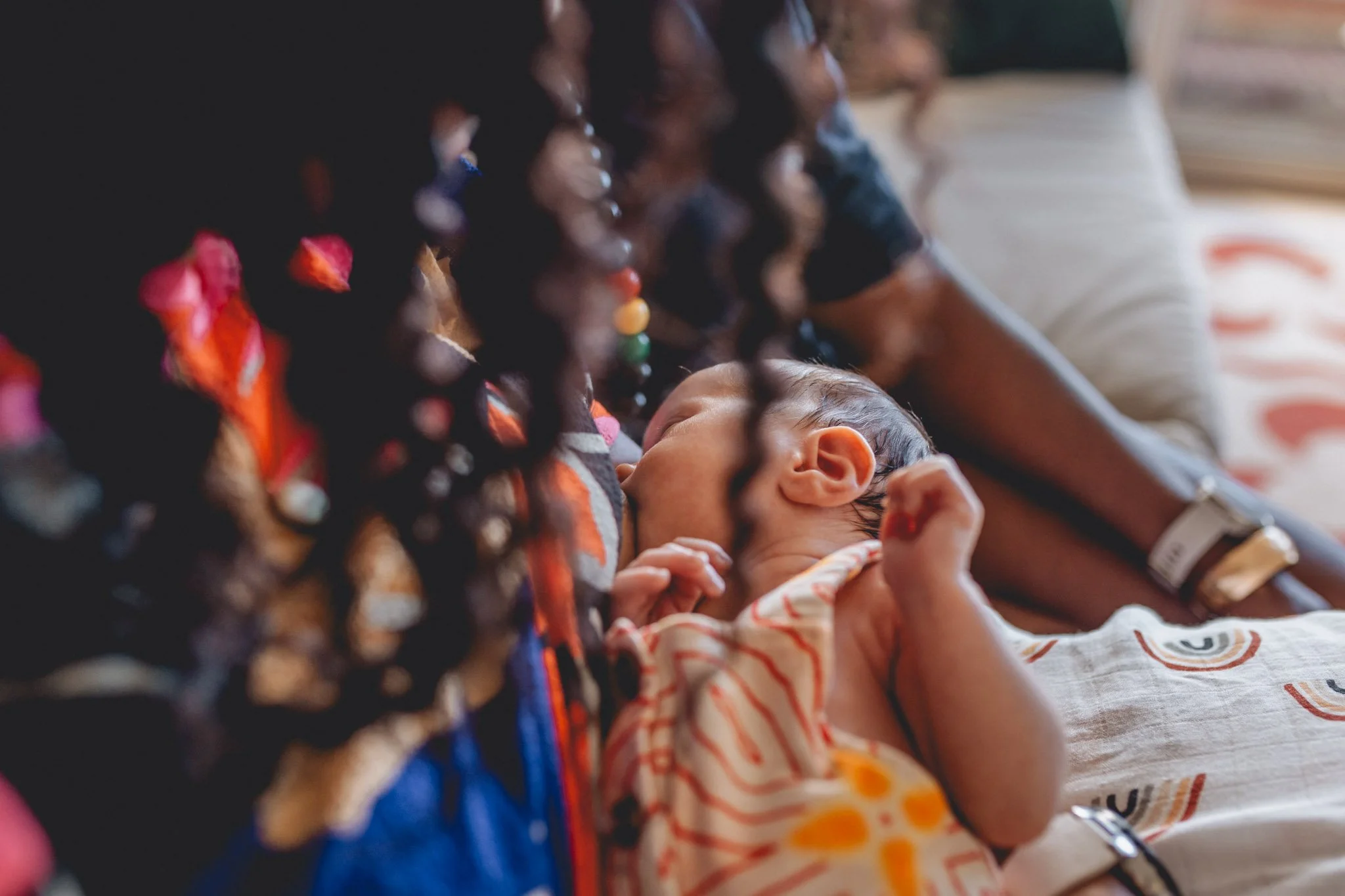 A person with dark curly hair and jewelry is holding a sleeping baby with a striped outfit, resting on the person's arm in a relaxed setting.