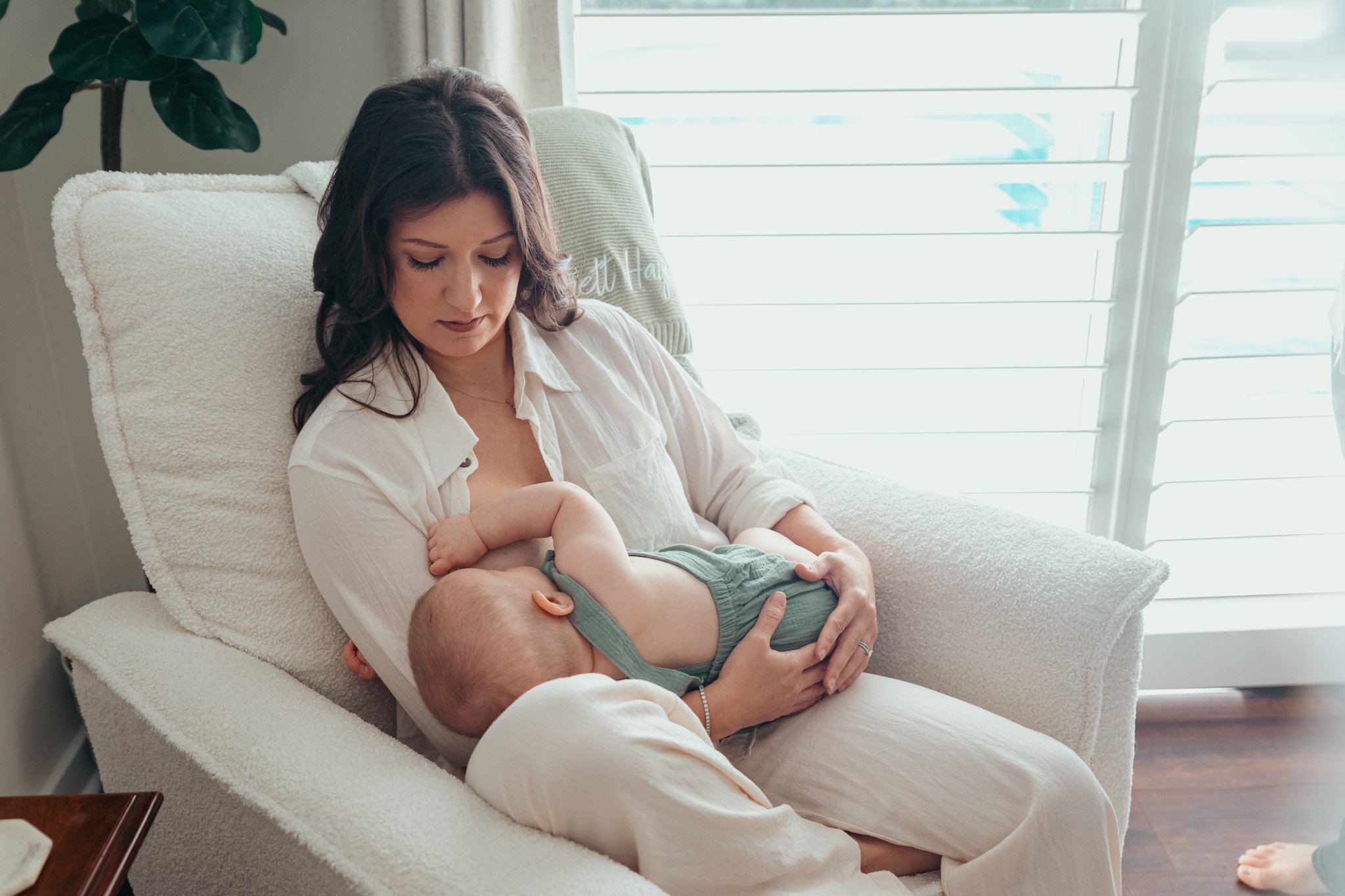 A woman with dark hair holding a baby while sitting on a cream-colored couch in front of a window with blinds.