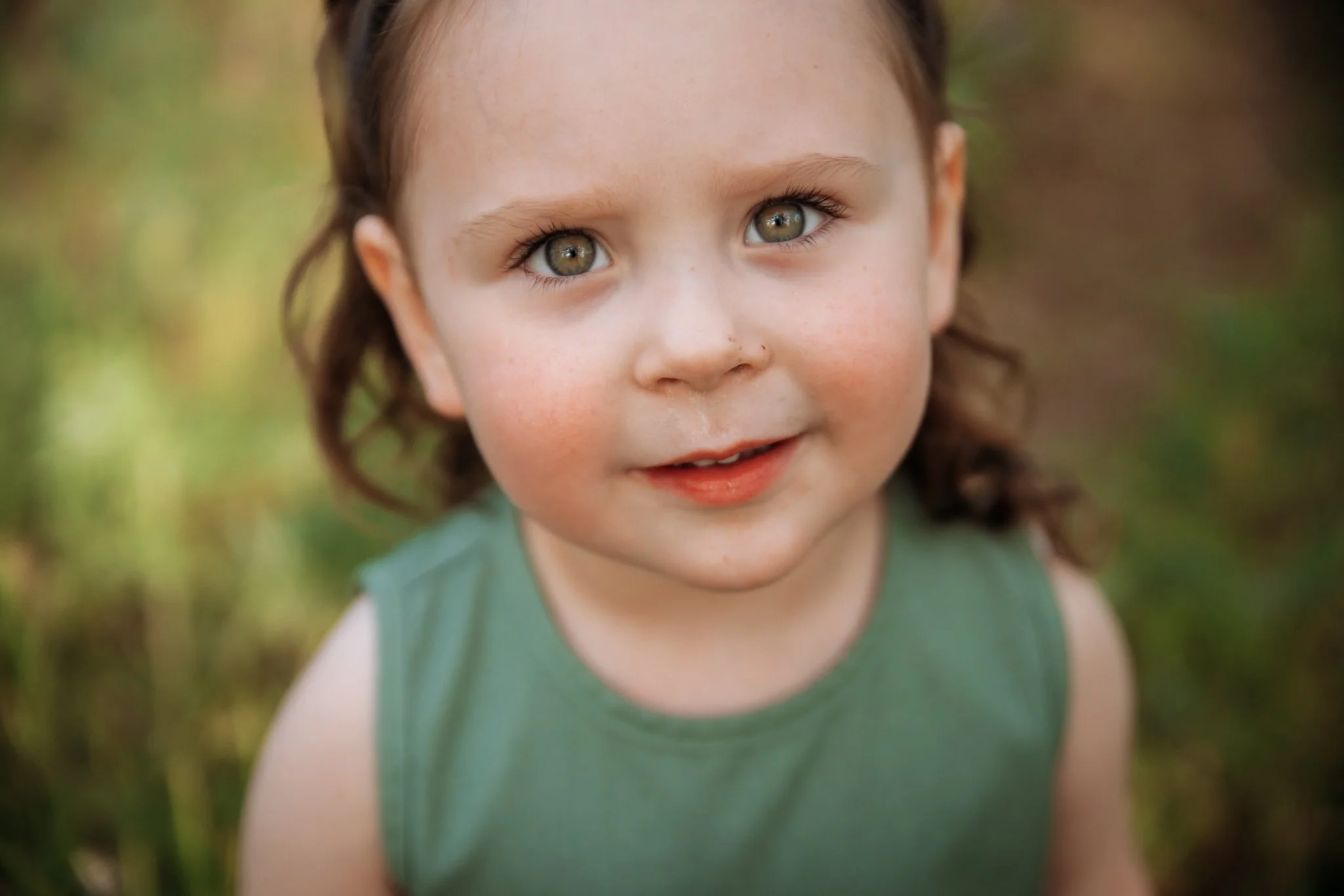 Close-up of a young girl with curly brown hair and hazel eyes, smiling outdoors with a blurred background of trees and grass.