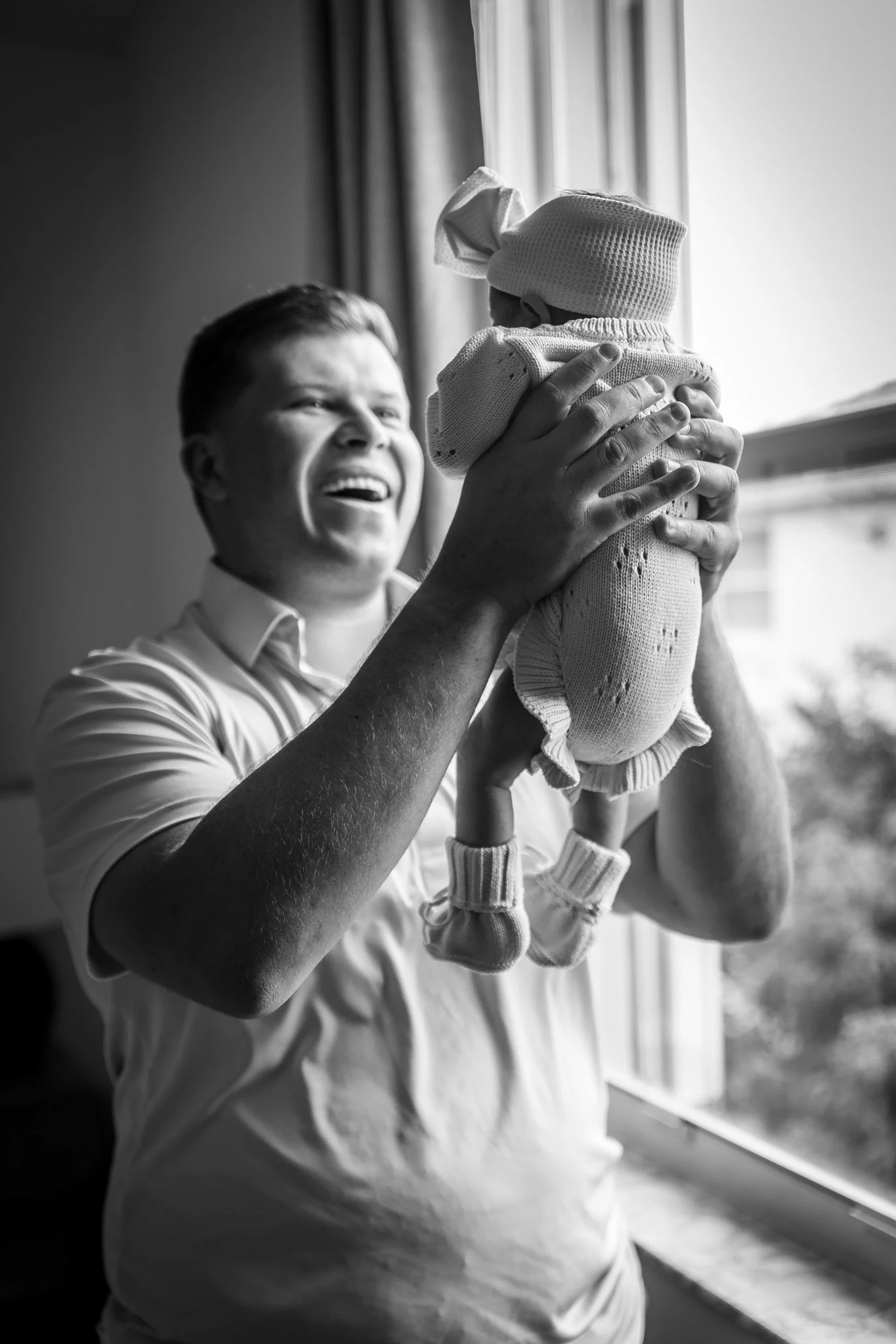 A man is holding a baby up near a window. The baby wears a hat and sweater, and the man appears to be happy, smiling while looking at the child.