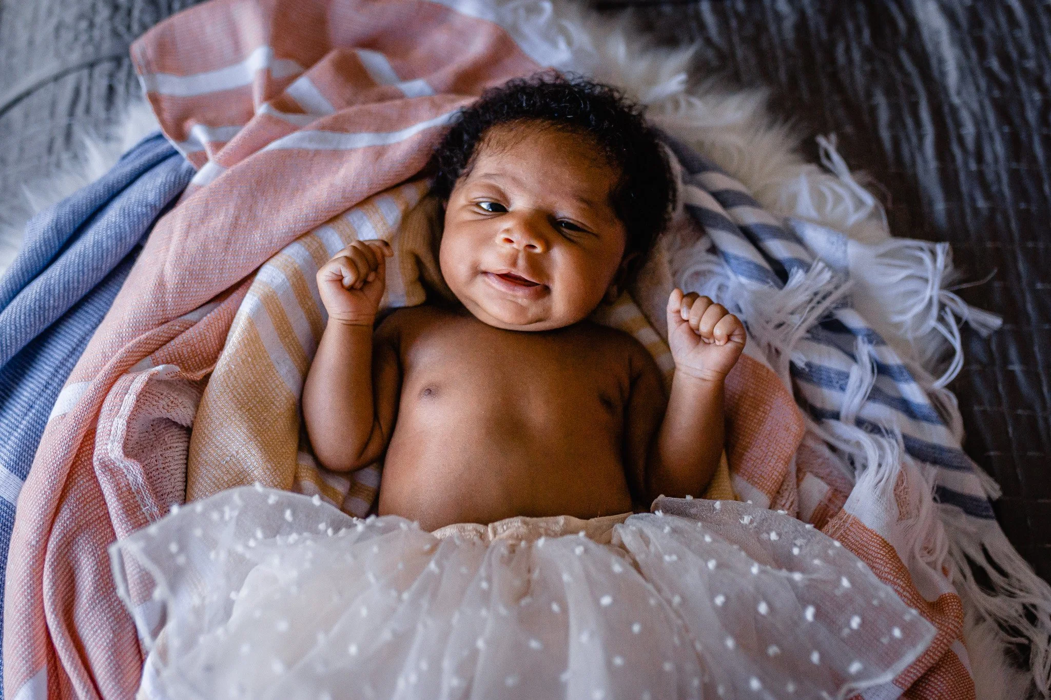 A smiling baby lying on a soft surface, partially covered with a blanket, with a protective paper in the foreground.