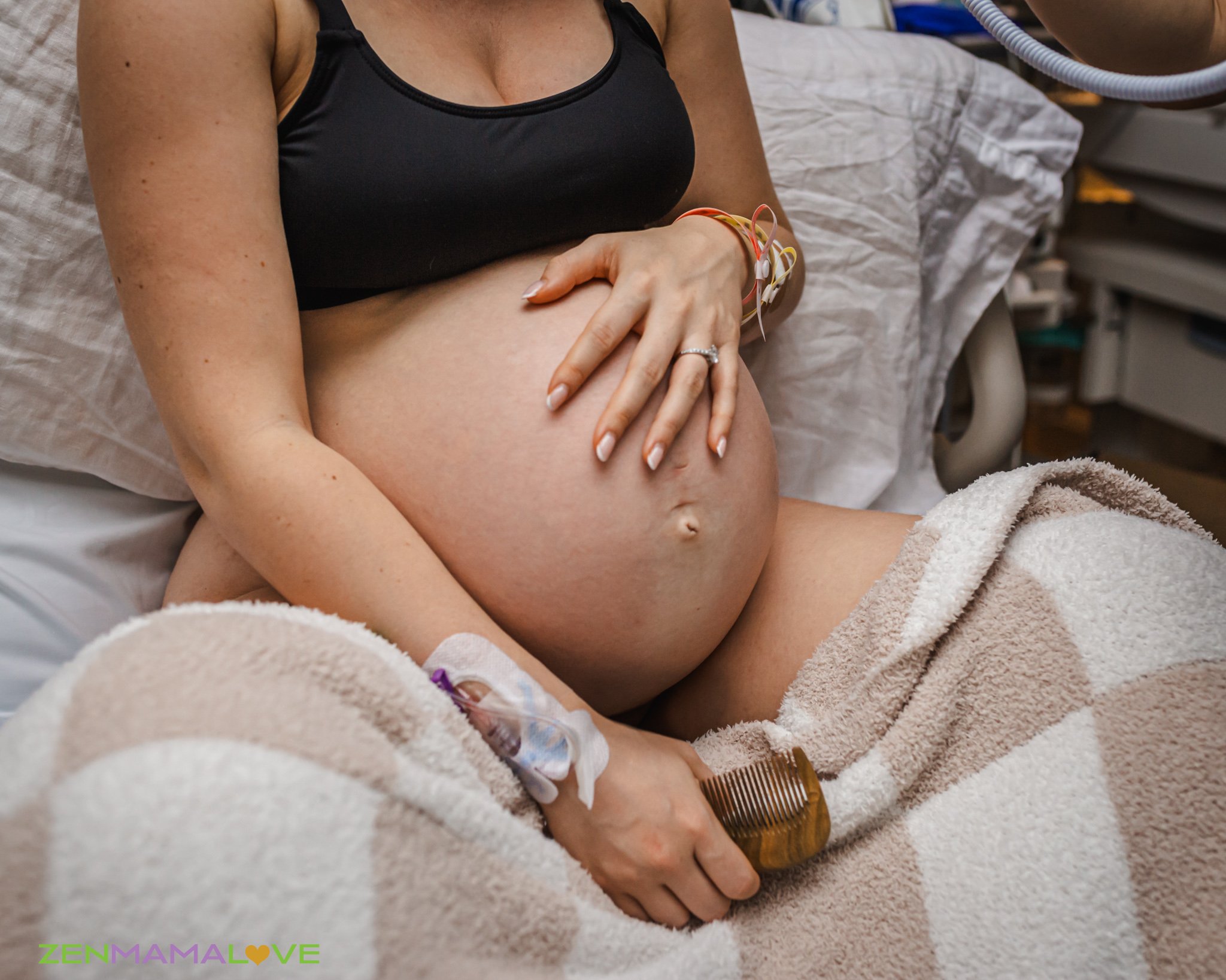 A woman lying in a hospital bed with medical monitoring wires on her arm, wearing a black sports bra, holding her pregnant belly with her hand, and a wooden comb is near her belly. The background shows medical equipment and a hospital bed.