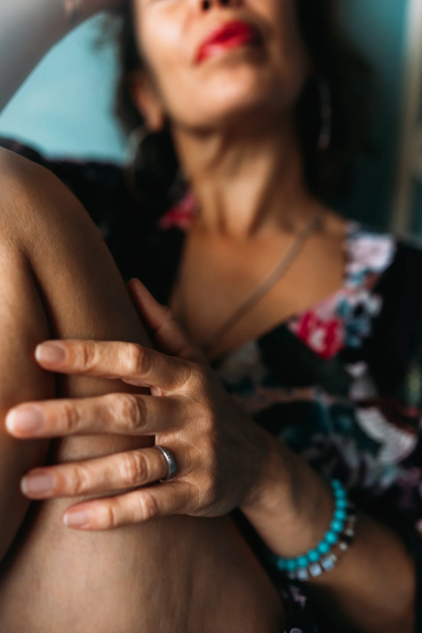 A woman is lying down with her face partially visible, wearing red lipstick, earrings, and necklaces. She is holding her arm with her hand, and the focus is on her arm and hand.