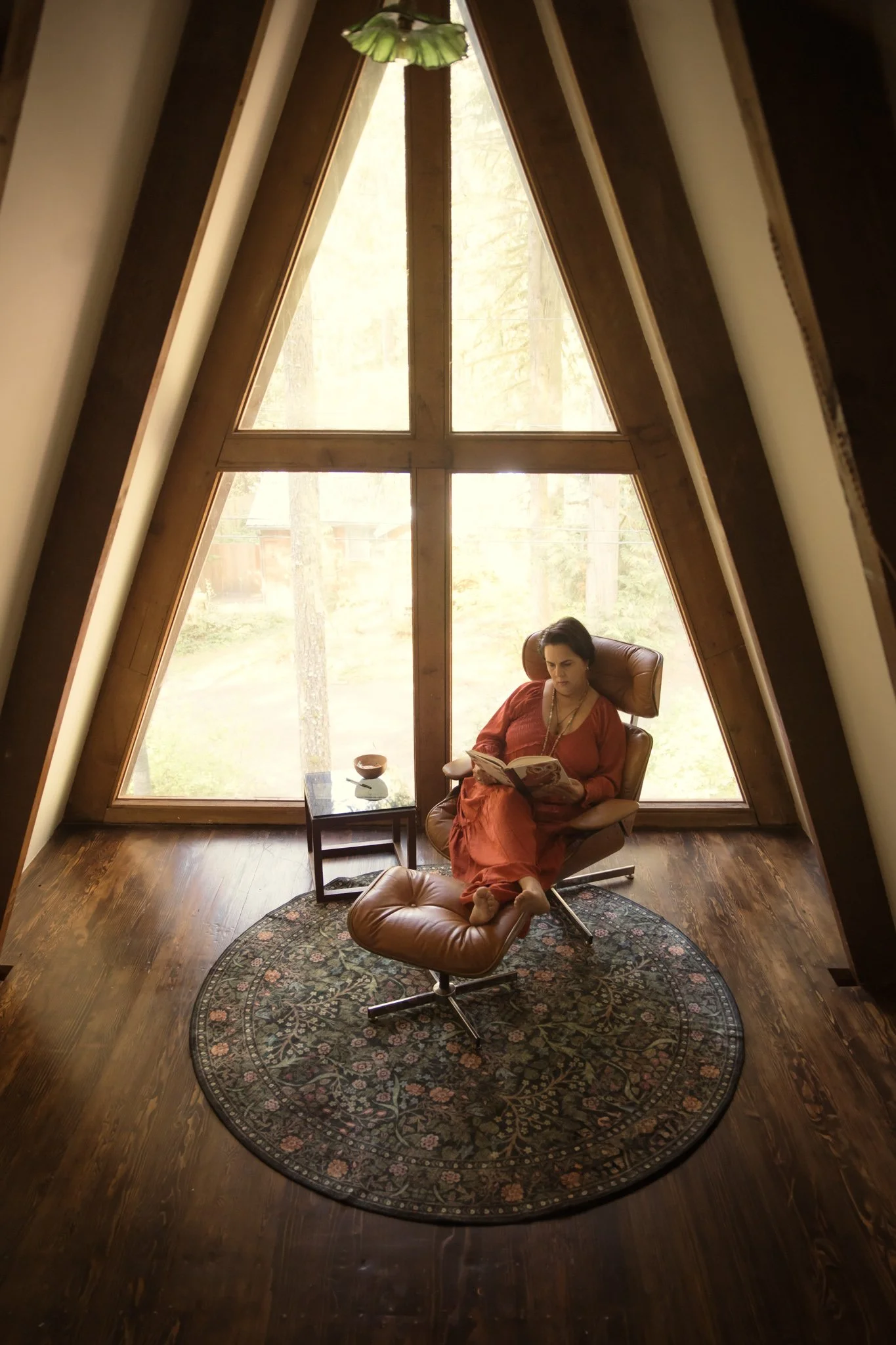 A woman sitting in a leather chair with a matching footrest, reading a book, in a room with large triangular windows and wooden framing, a green ceiling light, and a patterned rug on wooden floor.