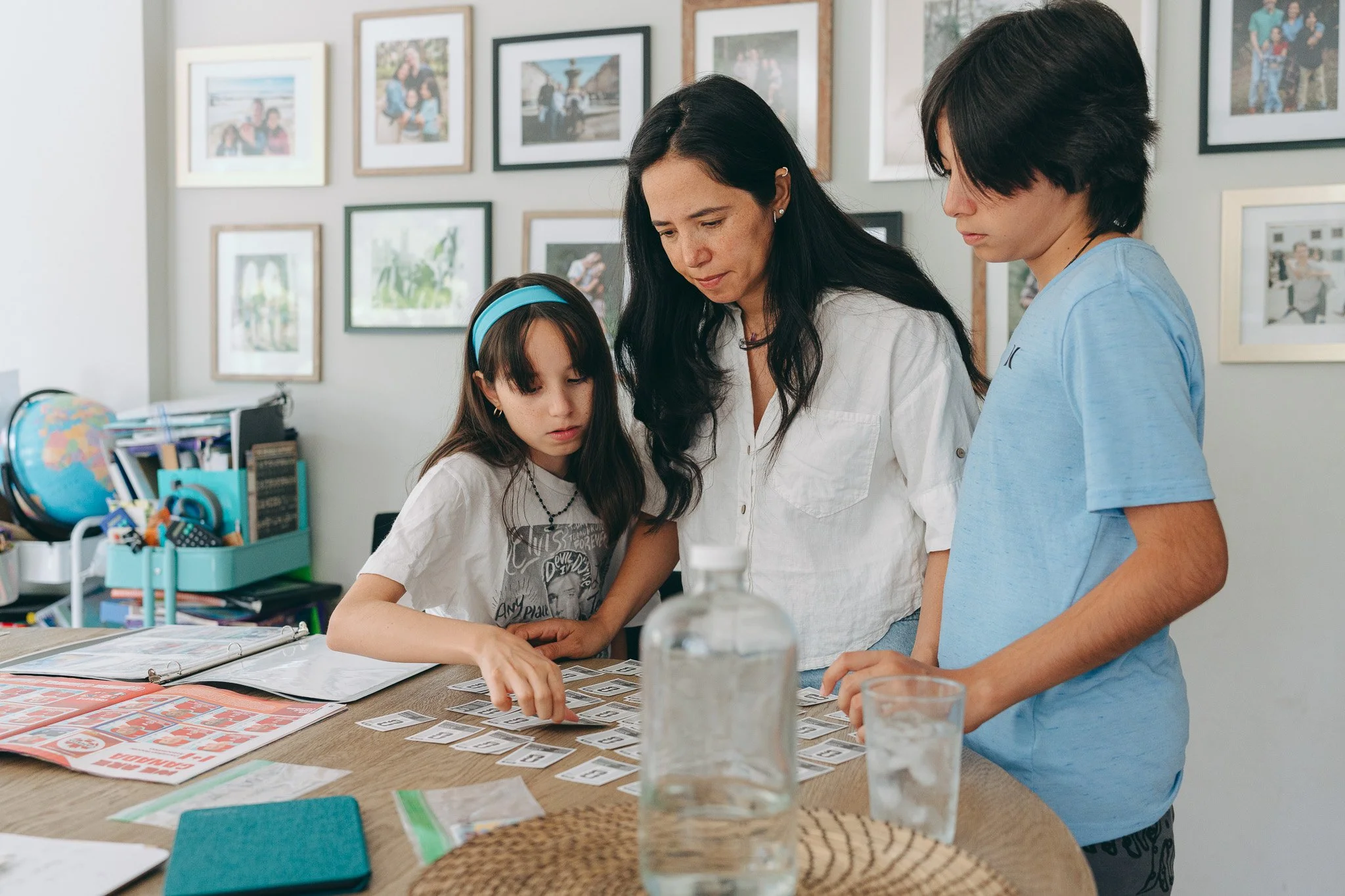 Three people, two women and a girl, are gathered around a table looking at cards. The girl is pointing at a card. The scene is in an indoor space with framed photos on the wall behind them. There are glasses of water and various papers or cards on th