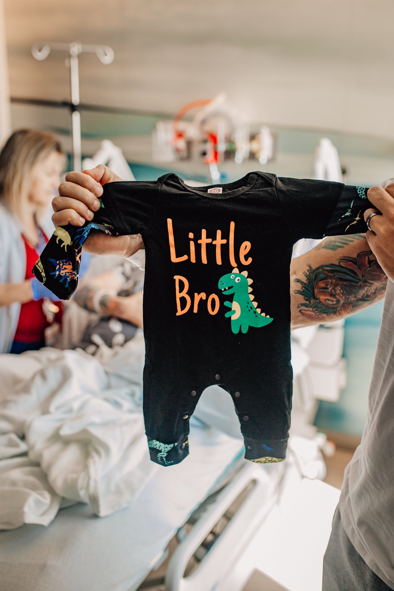 A person holding up a black baby onesie with colorful dinosaur design and the words 'Little Bro' in a hospital room.