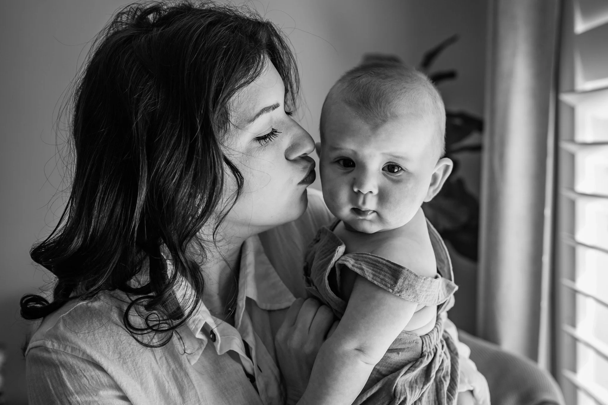 A woman holding and gently kissing a young child's head inside a house, near a window with blinds.