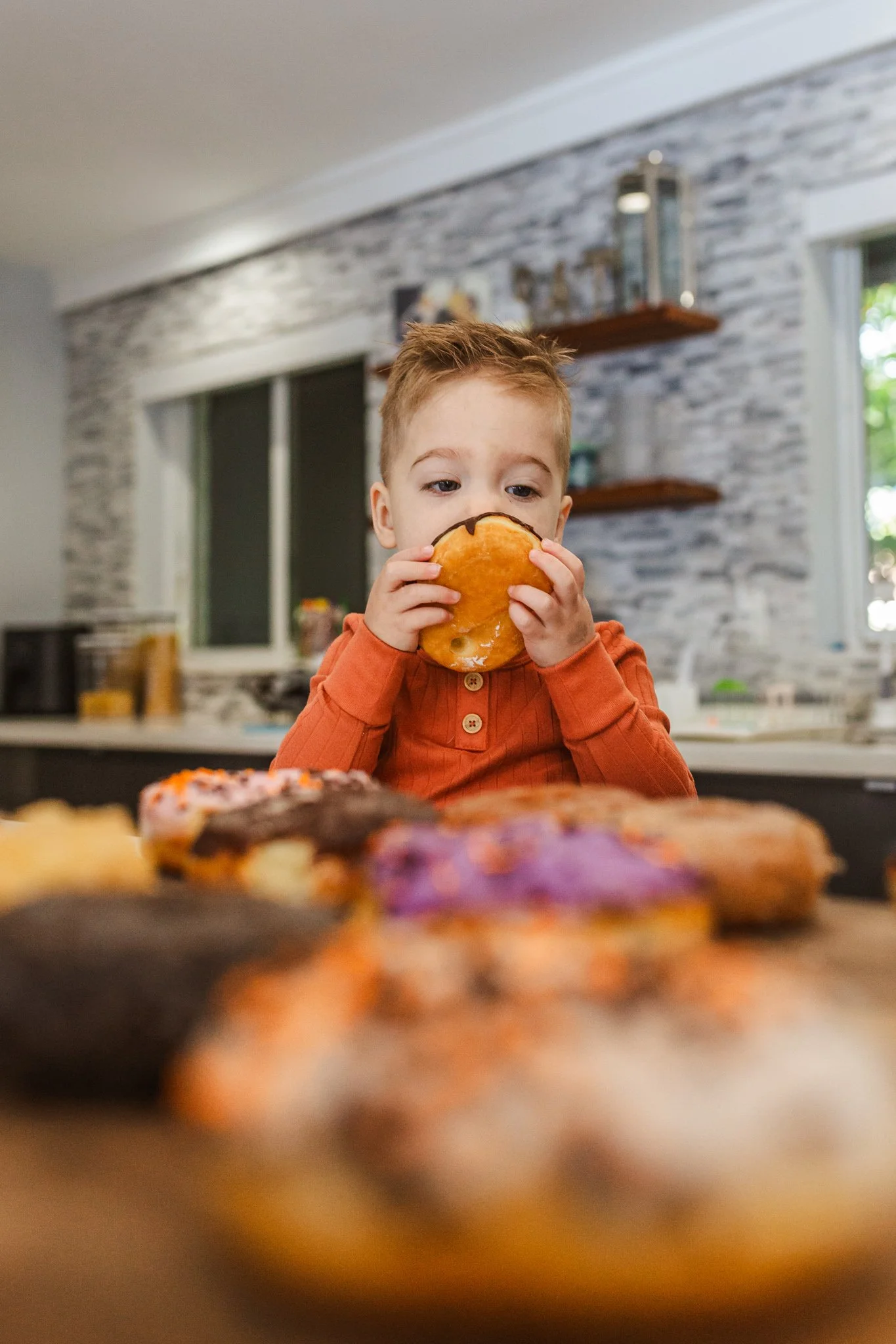 A young boy with short, light brown hair wearing an orange shirt is tasting a glazed donut while standing in a kitchen. There are colorful donuts on the table in the foreground and a kitchen with shelves and a window in the background.