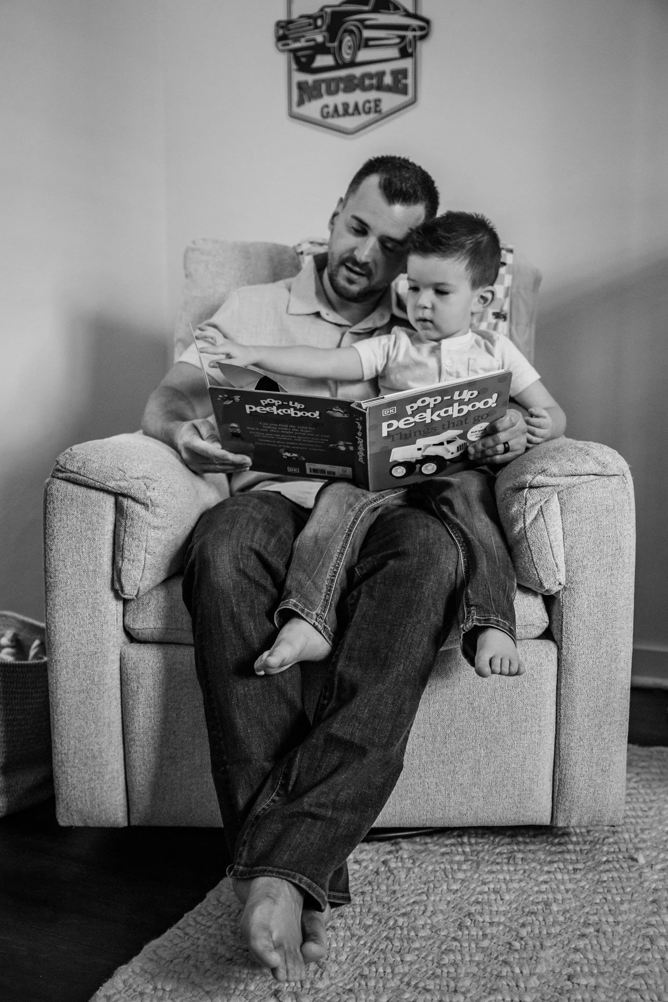 A man and a young girl sitting on a couch. The man is reading a book titled 'peekaboo! Things that go' while the girl reaches out, touching the book. There is a sign on the wall that says 'Muscle Garage' with a car illustration. The scene is in black