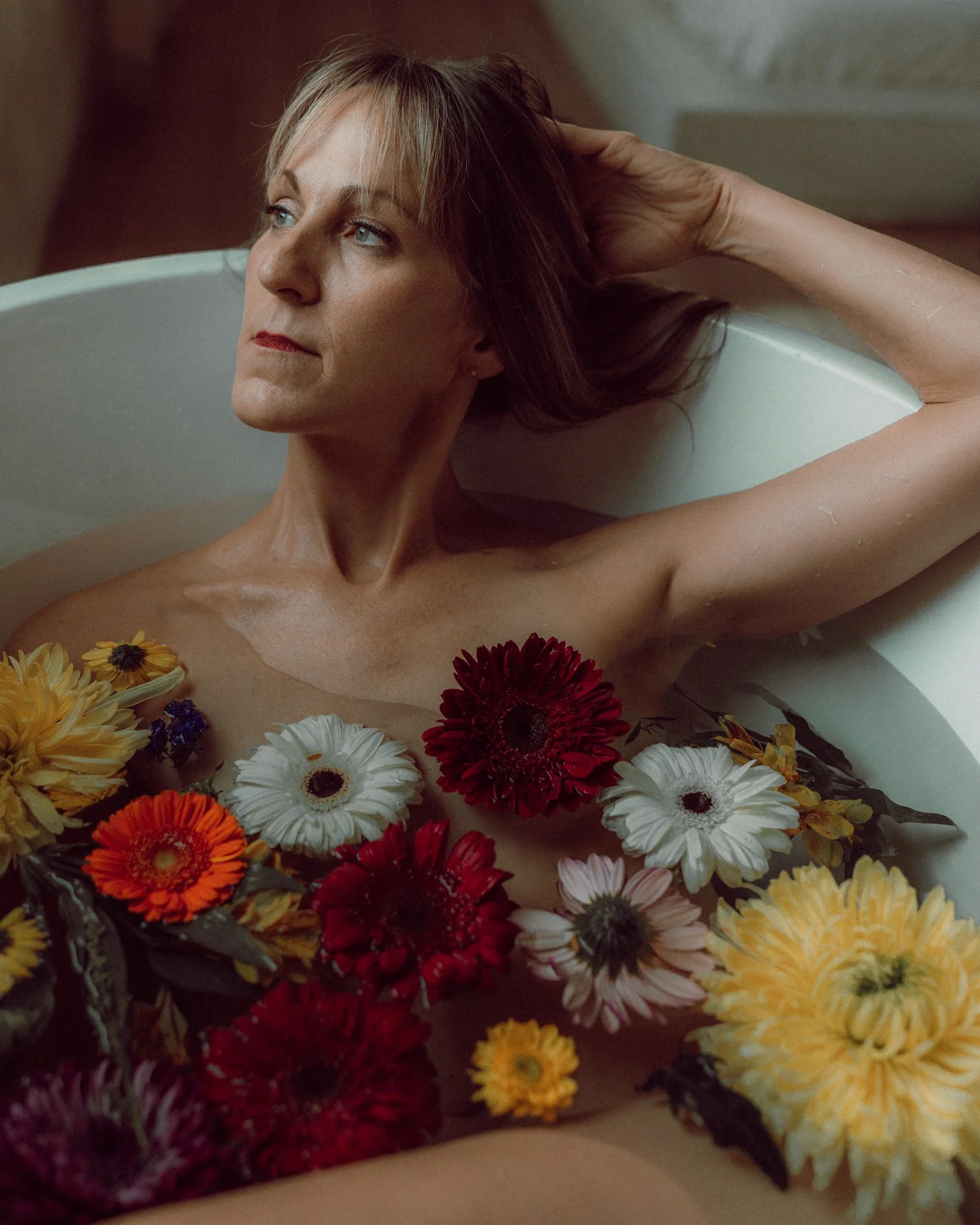 A woman with reddish hair relaxing in a bathtub surrounded by colorful flowers including yellows, oranges, reds, and whites.