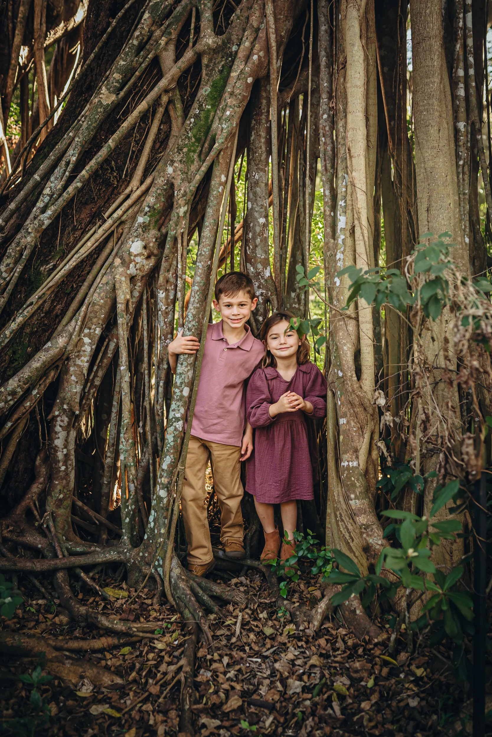 Two children, a boy and a girl, standing inside the large roots of a tree in a forest, smiling at the camera.