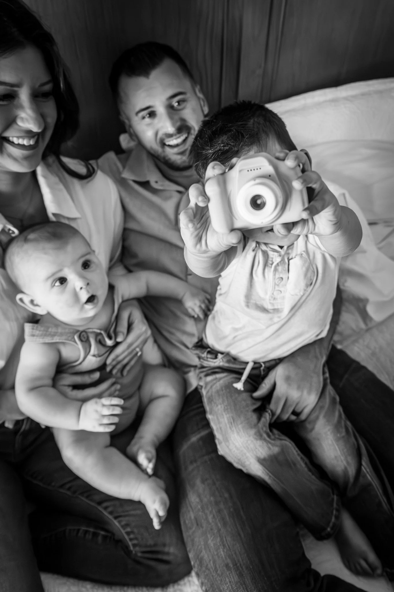A family of four sitting together on a bed. The mother is smiling, a young man is smiling, a young boy is holding a camera and taking a picture, and a baby is sitting on the bed looking at the camera. The photo is in black and white.