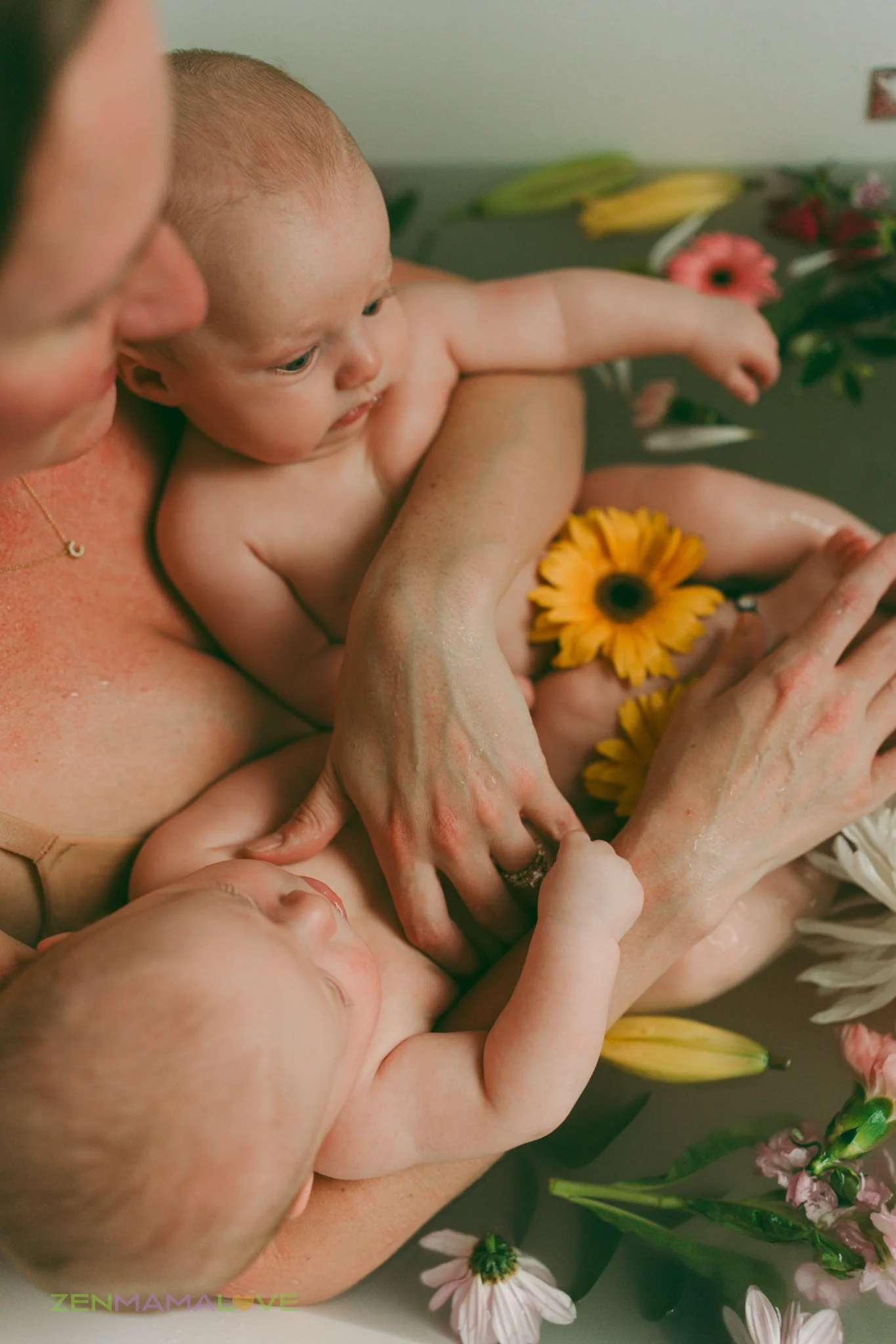 A newborn baby being bathed in a tub filled with water and colorful flowers, with a woman holding the baby, surrounded by additional flowers.