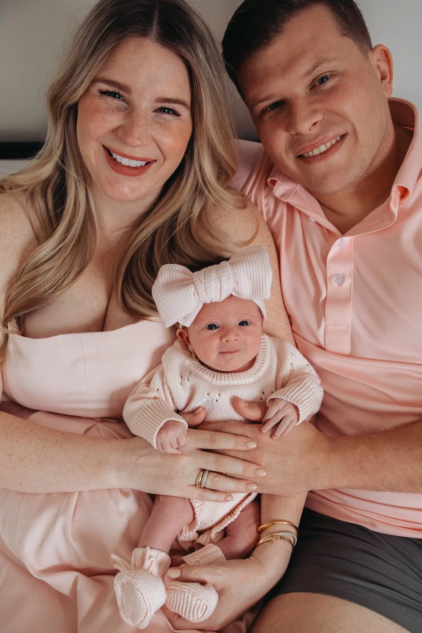 A family of three: a woman, a man, and a baby, smiling and sitting closely together. The woman has long, wavy light brown hair, freckles, and is dressed in a light pink or beige outfit. The man has short brown hair, a light complexion, and is wearing