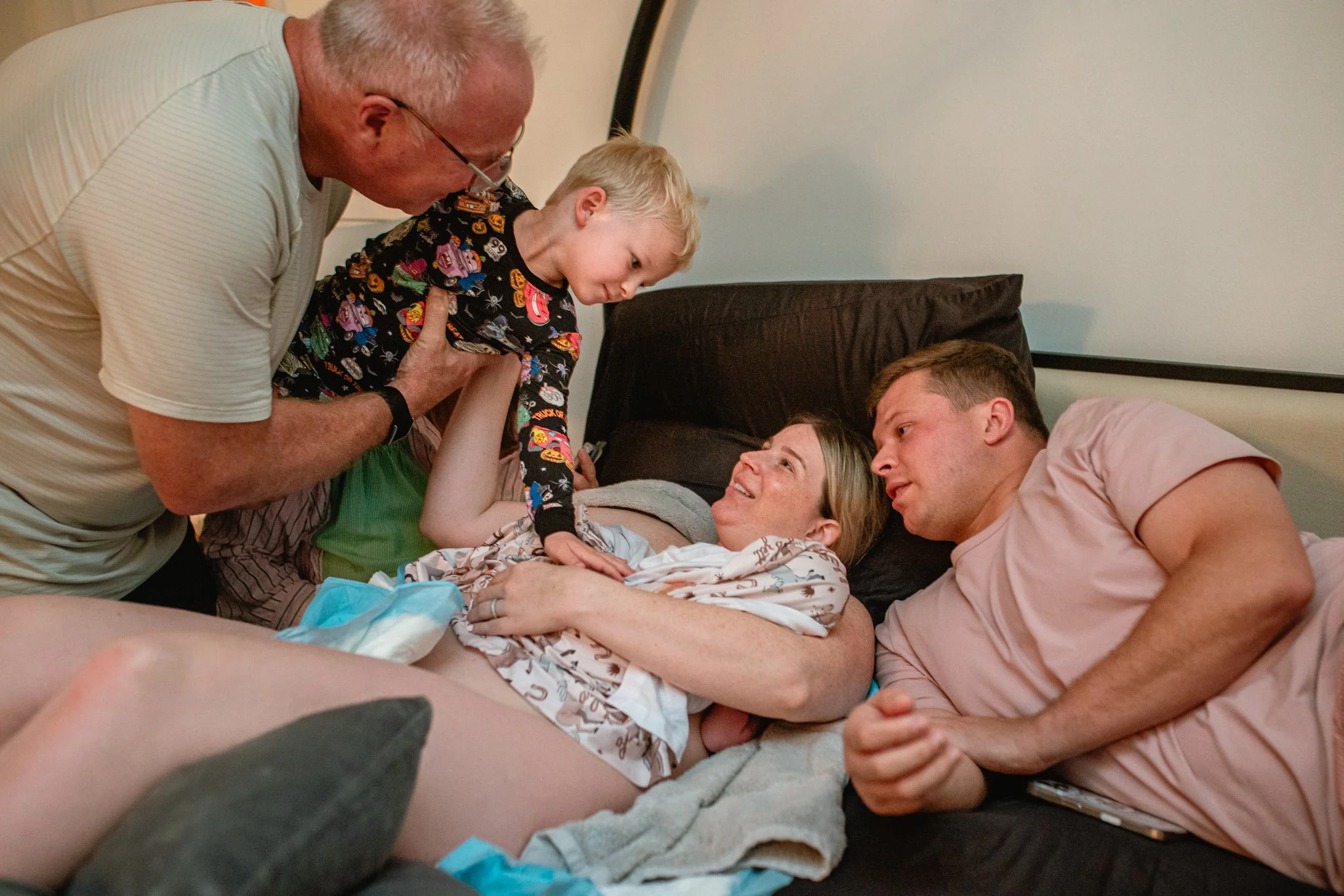 Family members gathered around a woman in a hospital bed, with a young boy sitting on the bed, a man lying beside her, and an older man leaning over, all engaging closely and lovingly.