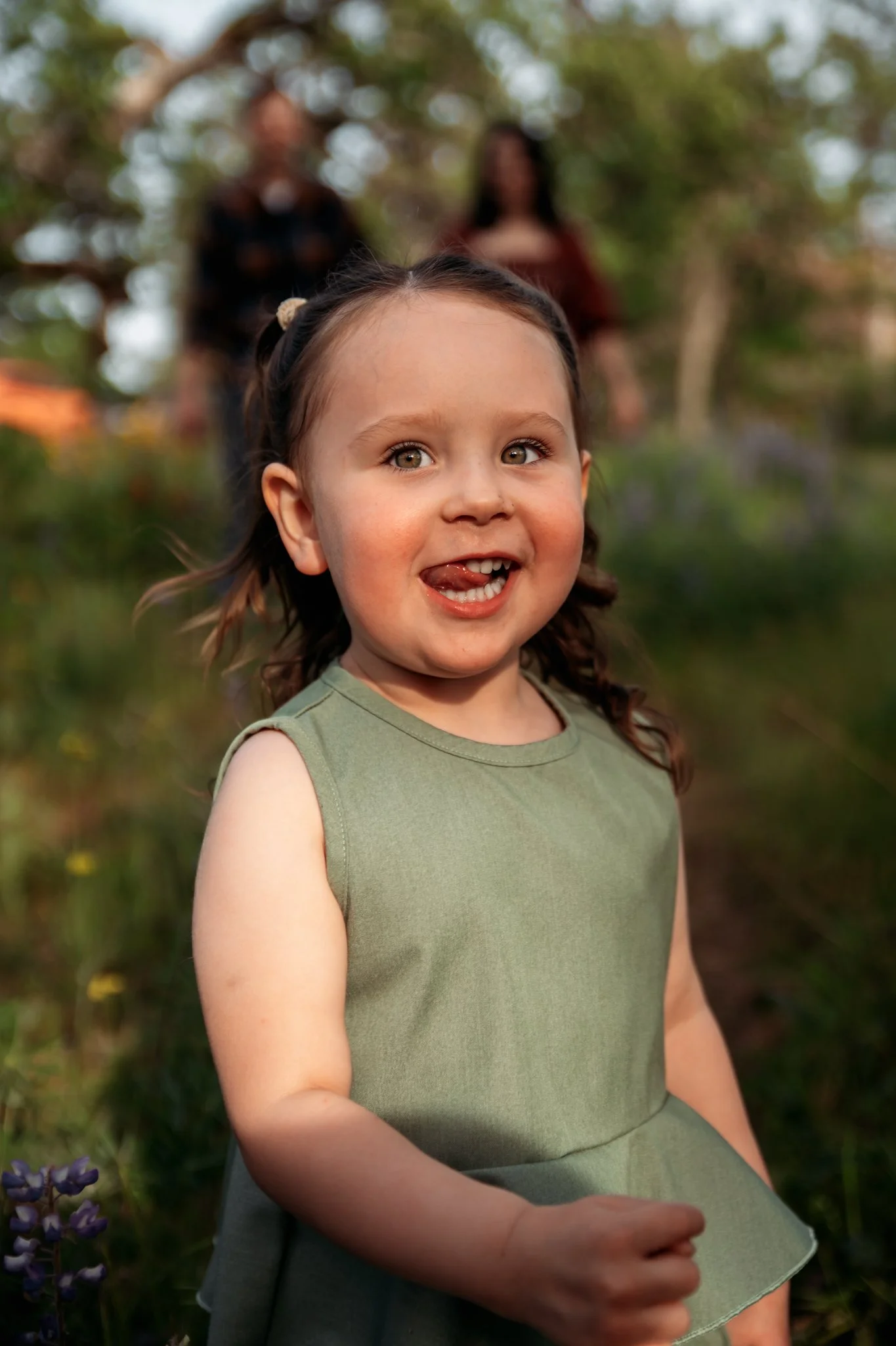 A young girl smiling outdoors with greenery and blurred two adults in the background.