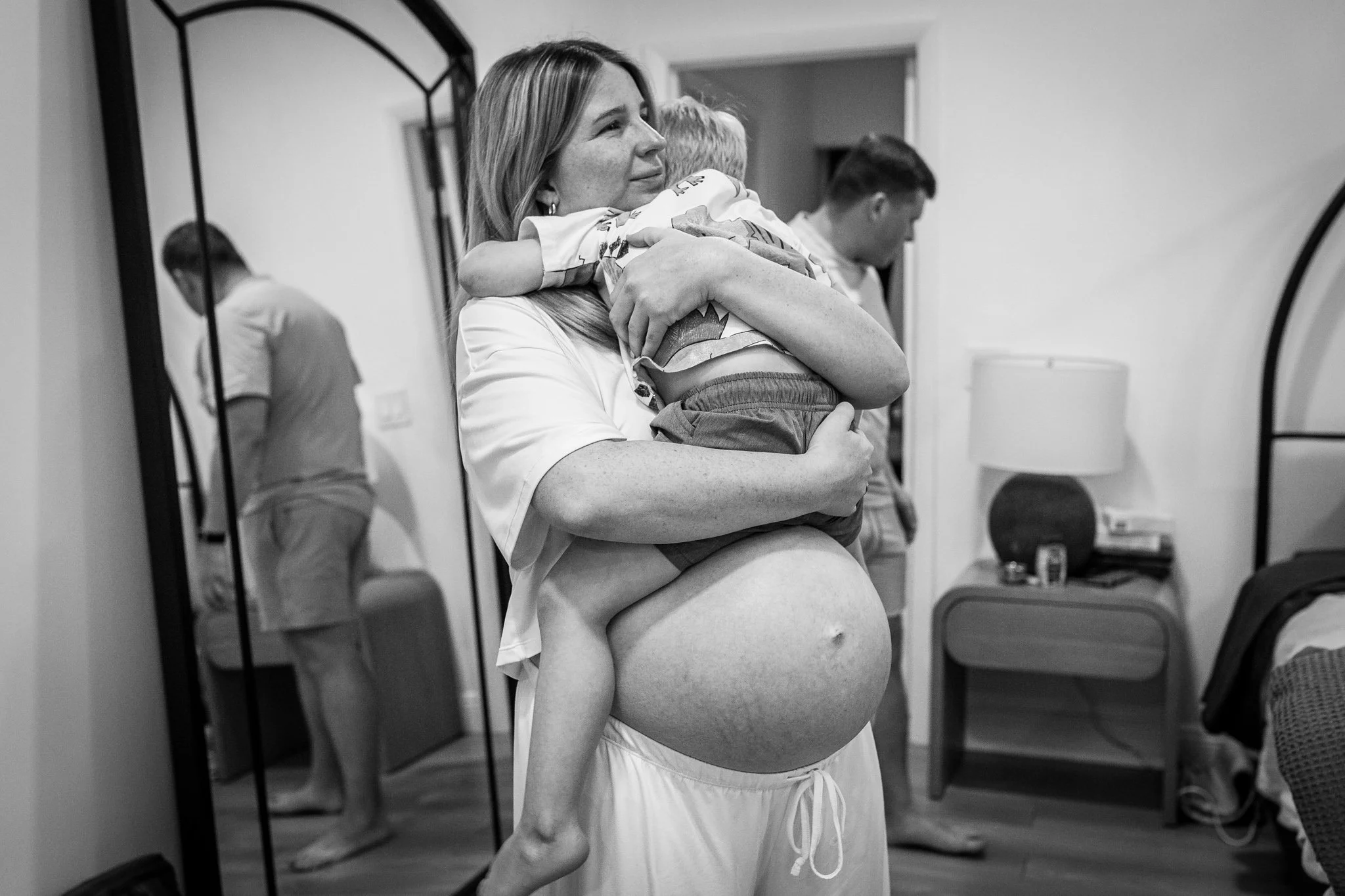 A pregnant woman is holding a young boy in her arms and hugging him close. They are standing in a room with a mirror, with a man and a boy in the background. The woman is smiling softly, and the scene appears warm and intimate.