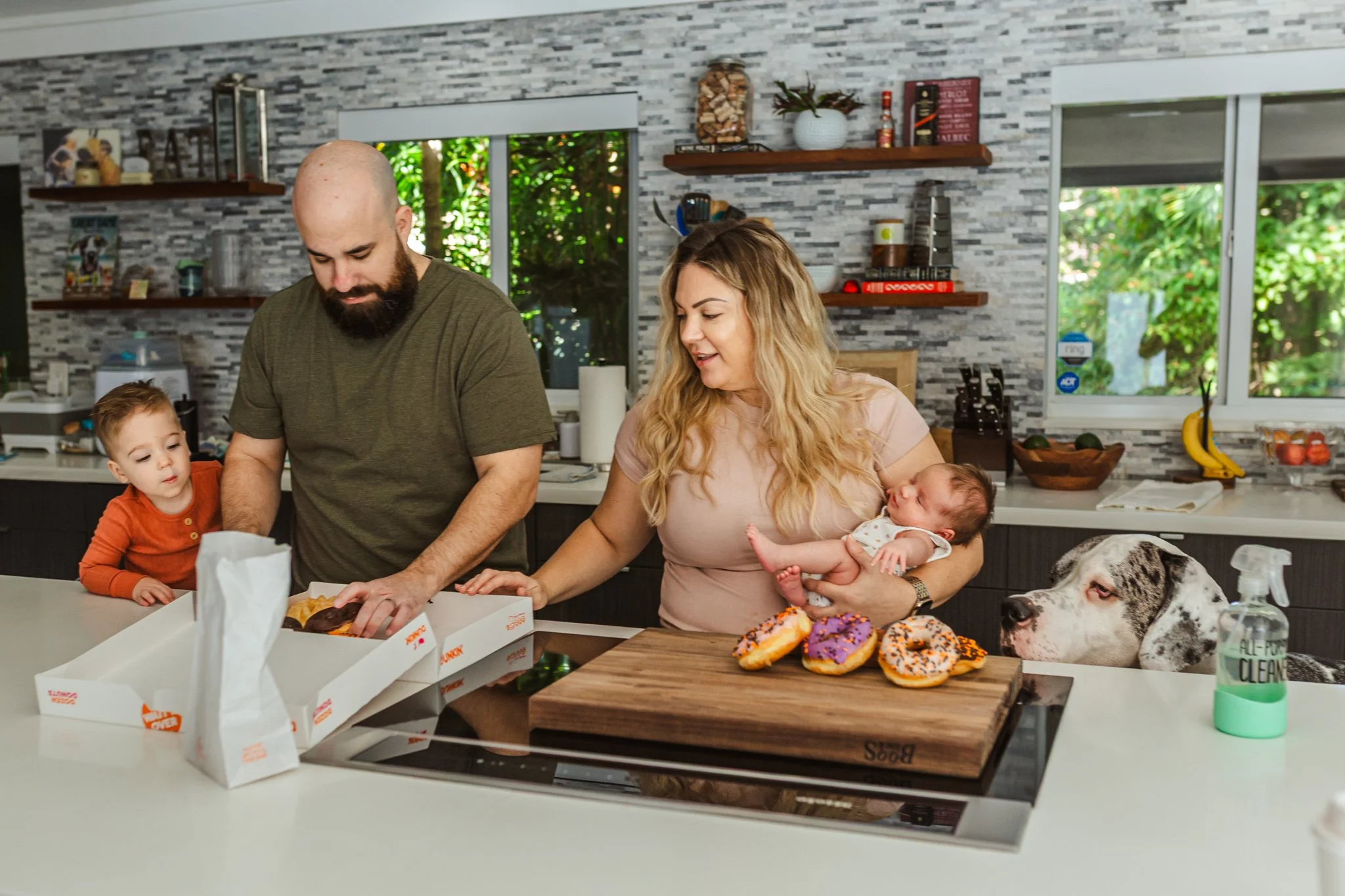 Family gathered in kitchen, with children and a woman holding a baby, preparing or serving food, donuts with purple icing and sprinkles on the counter, a man assisting, and a large dog resting nearby.