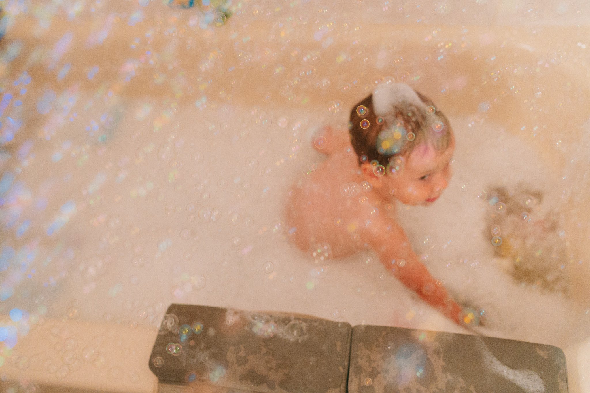A young girl with wet hair sitting in a bathtub filled with soapy water and glitter, smiling and looking to the side.