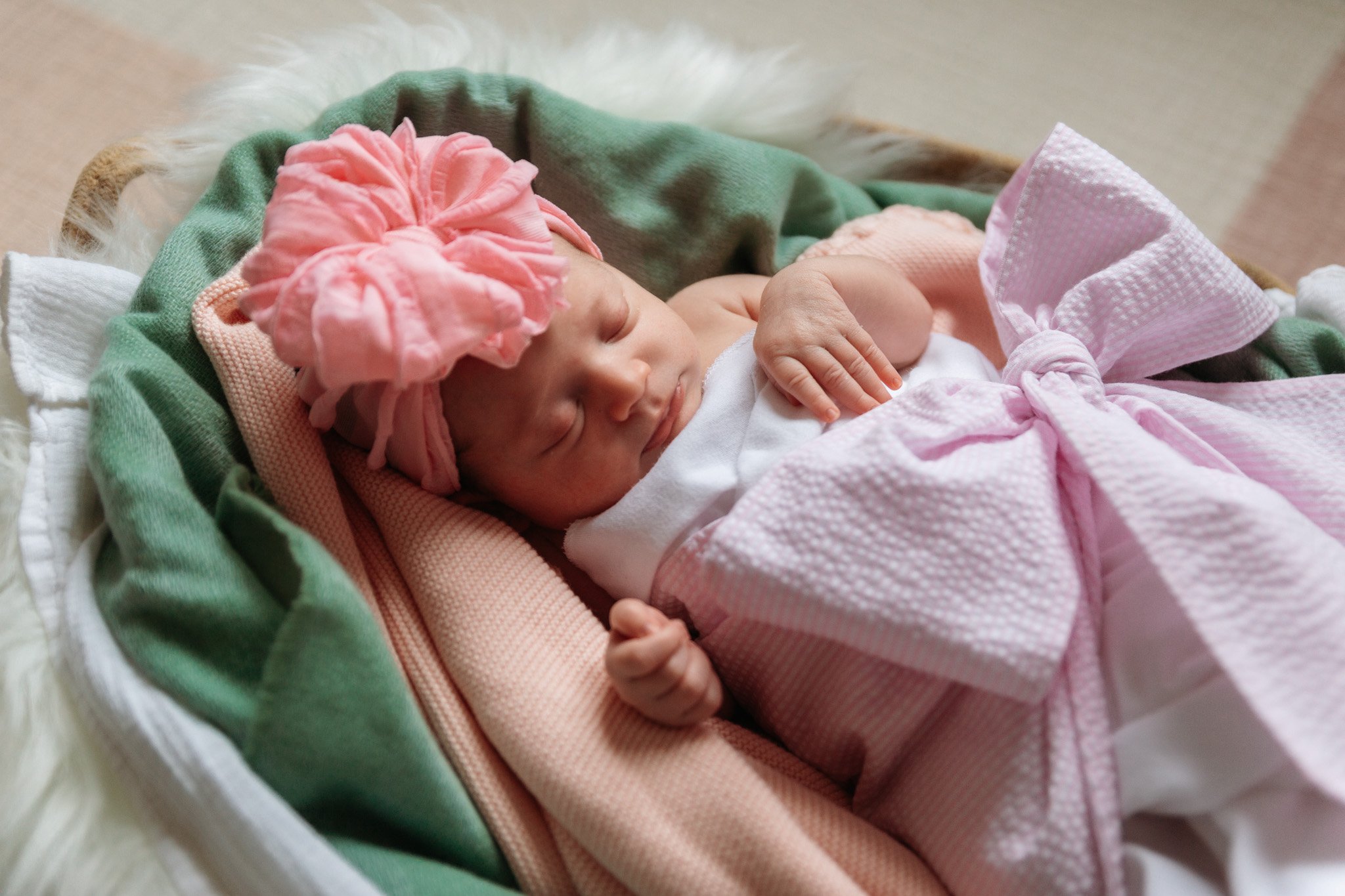A sleeping baby wearing a pink headband with a large pink flower and pink clothing, lying in a white basket lined with green and pink blankets.