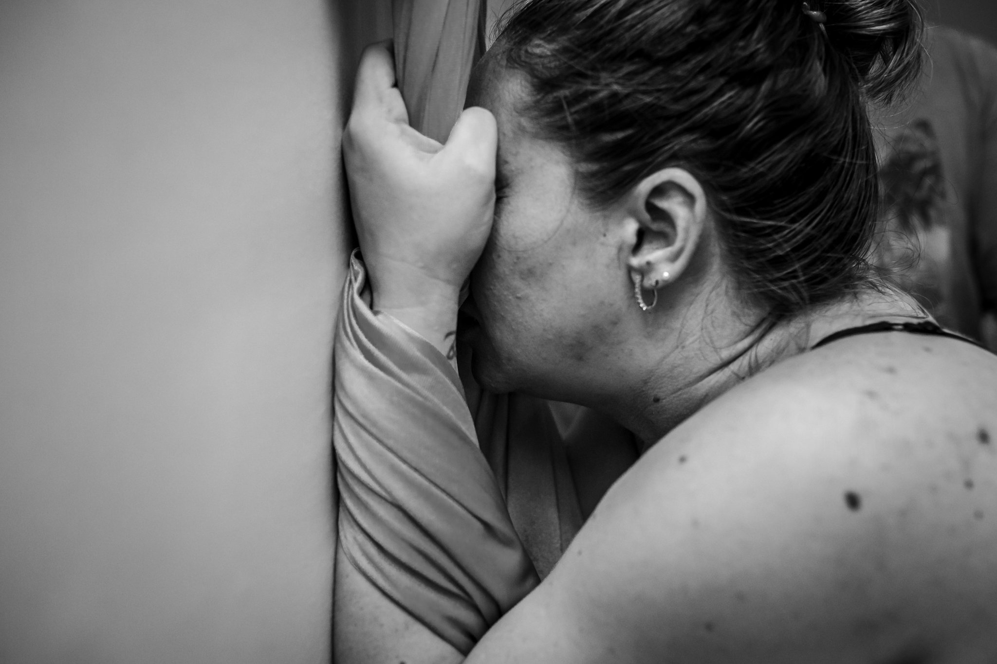 A woman with short hair and earrings leans against a wall with her hand on her head, expressing distress or contemplation in black and white.