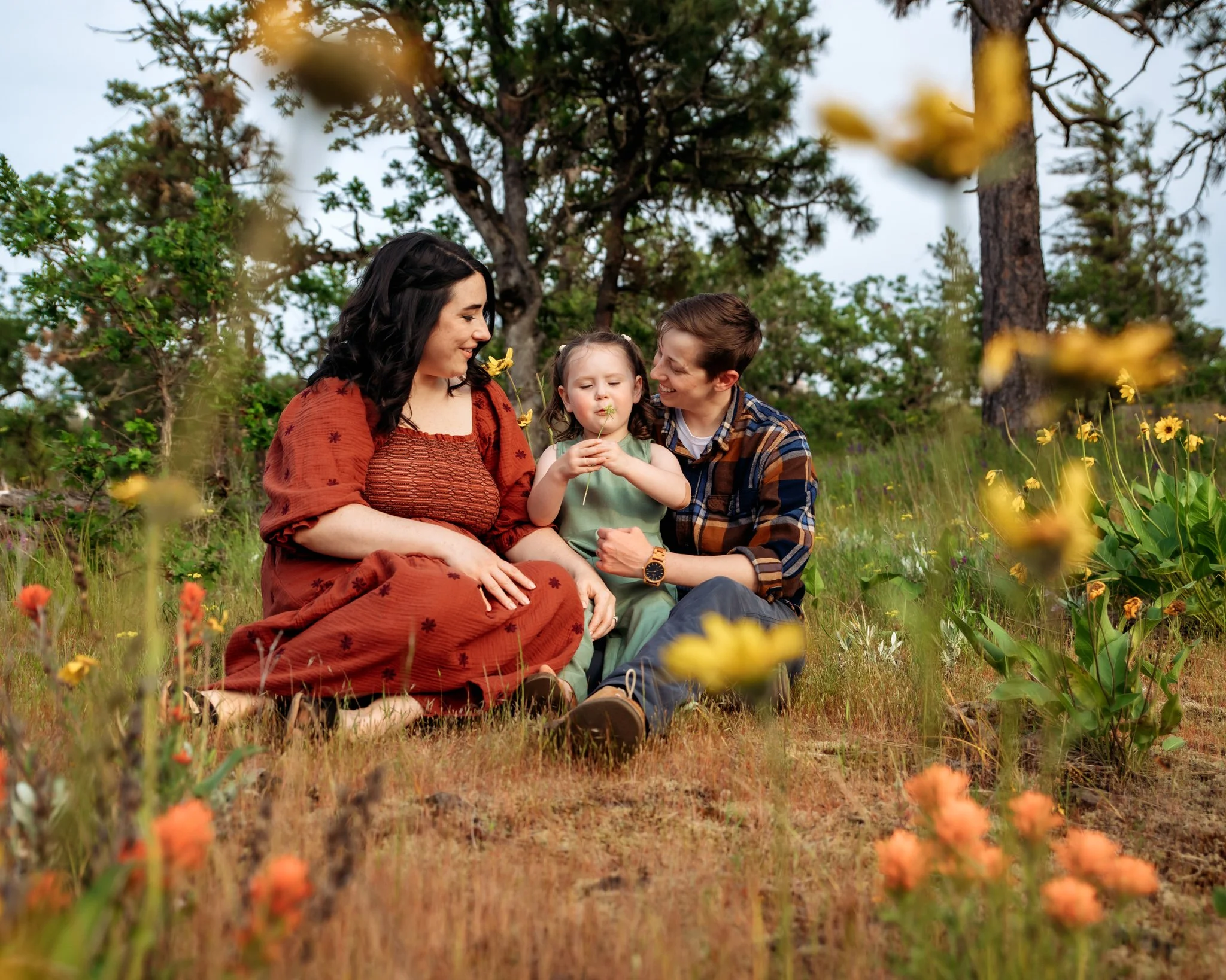 A family of four sitting outdoors in a grassy area with yellow flowers, trees, and blue sky in the background, enjoying nature and each other.