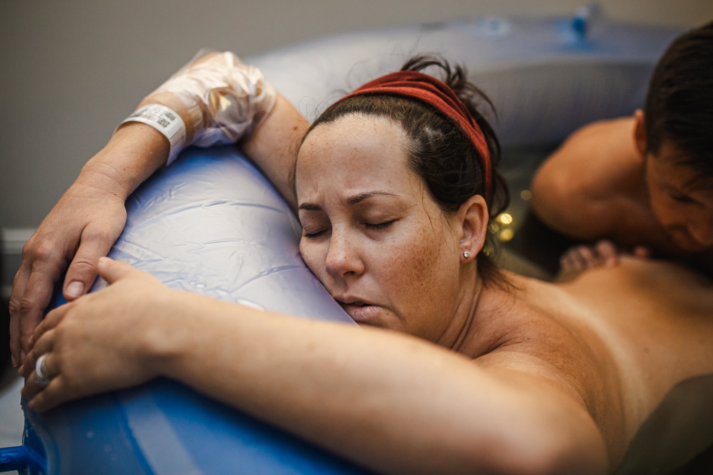 A woman with freckles and a red headband hugging a blue exercise ball with her eyes closed, while a man is beside her.