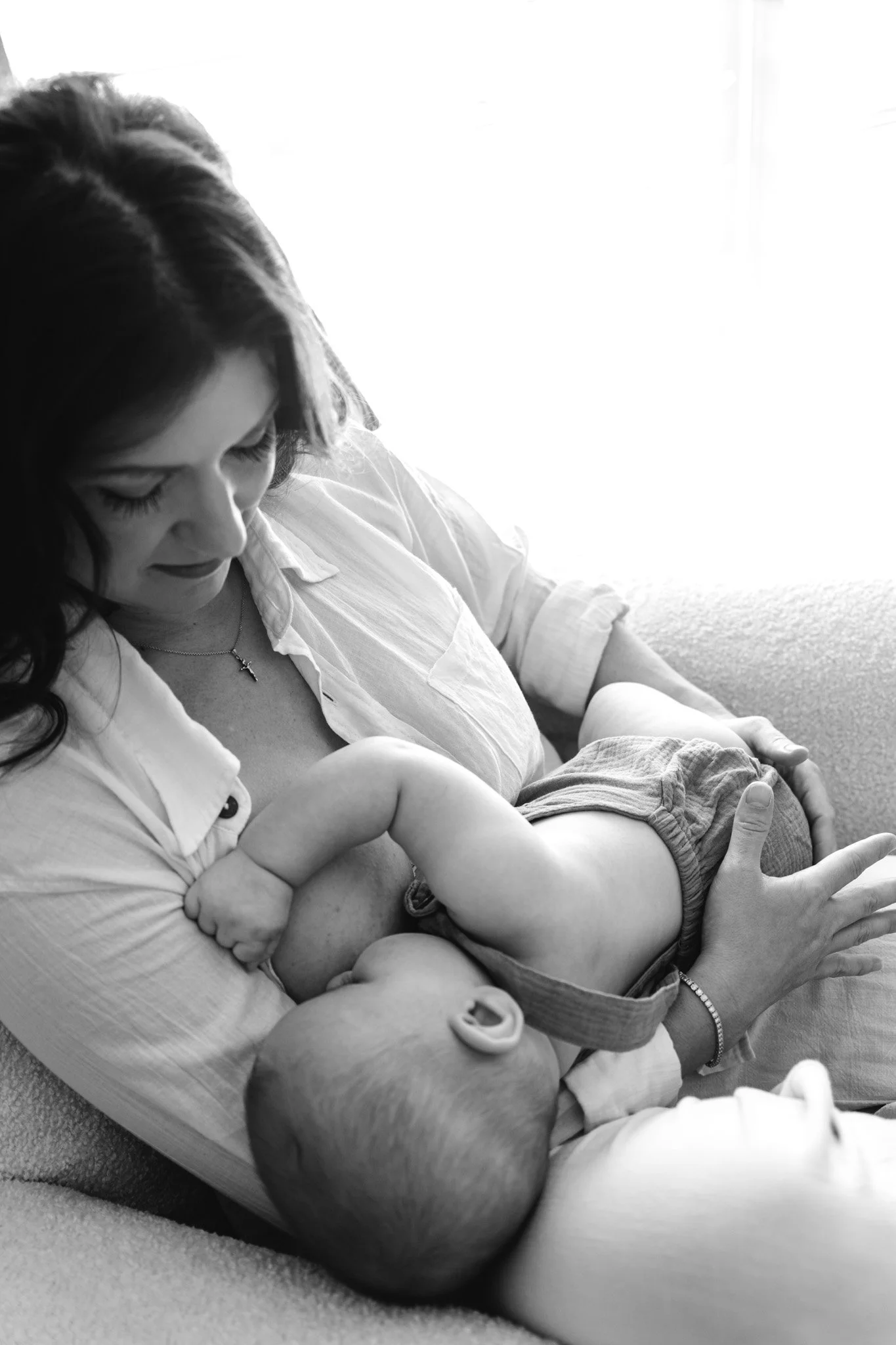 A woman holding and looking at a baby, both sitting on a soft surface. The woman is wearing a white shirt with rolled-up sleeves and a necklace. The baby is wearing shorts and a bracelet. The scene is bright with light coming from the background.