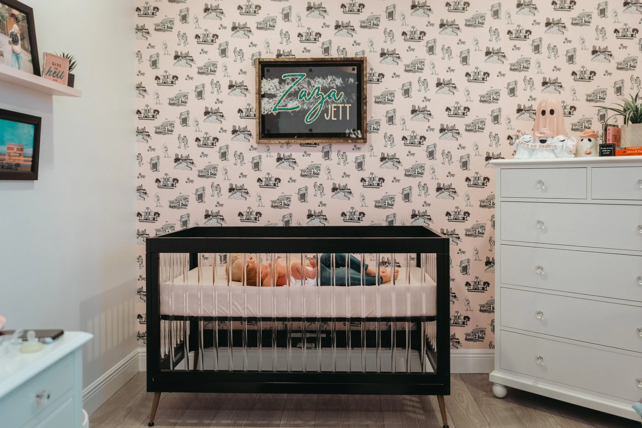A nursery with a baby lying in a crib, decorated with patterned wallpaper featuring animals and buildings. There is a framed sign on the wall that reads 'Zaza Jett'. To the right, a white dresser with decorative items, including a Halloween-themed co