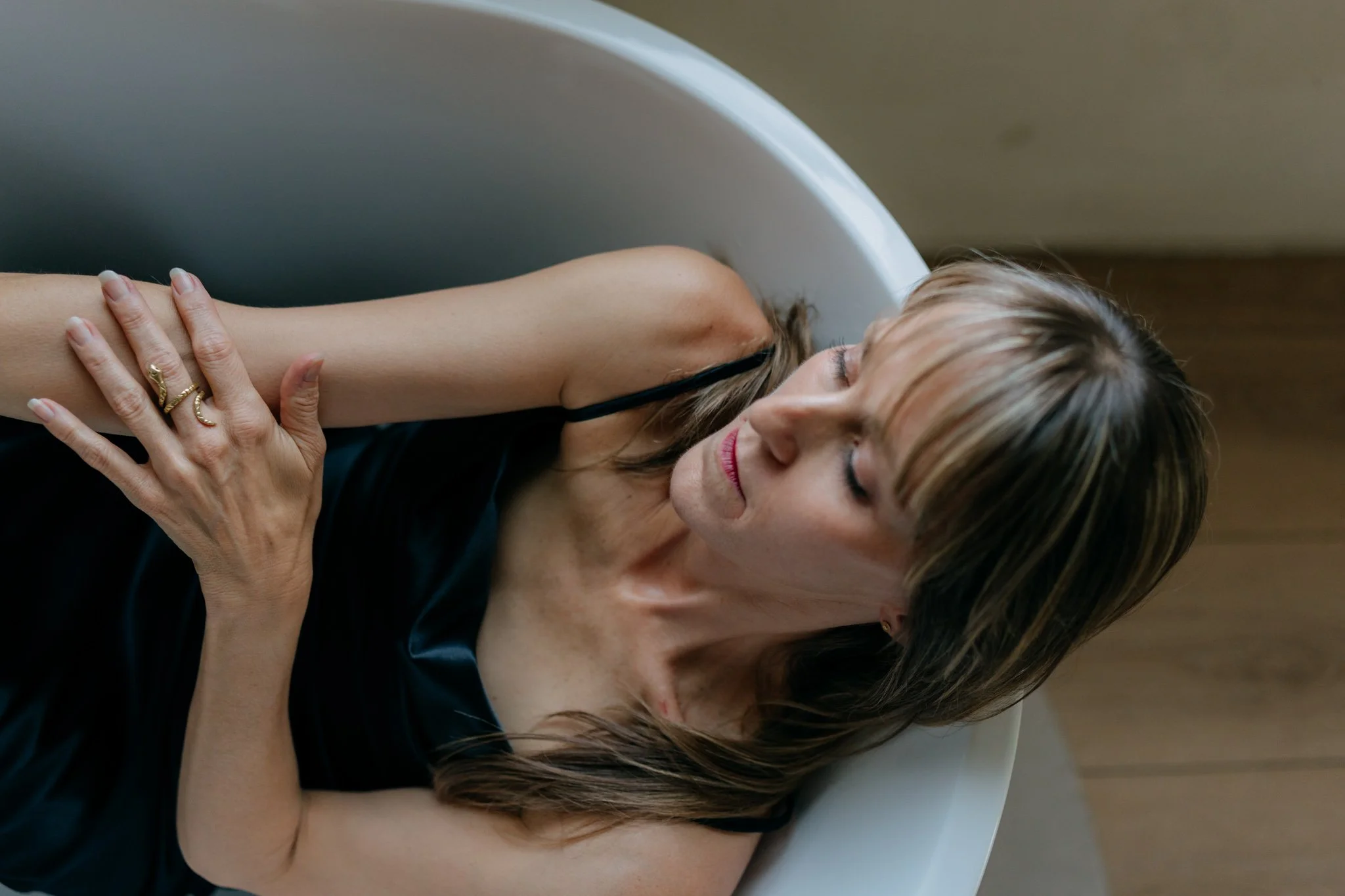 A woman with blonde hair and light skin is leaning into a bathroom sink, resting her head on the edge and closing her eyes. She is wearing a black sleeveless top and a gold ring on her left hand, which is resting on her arm. The background is neutral