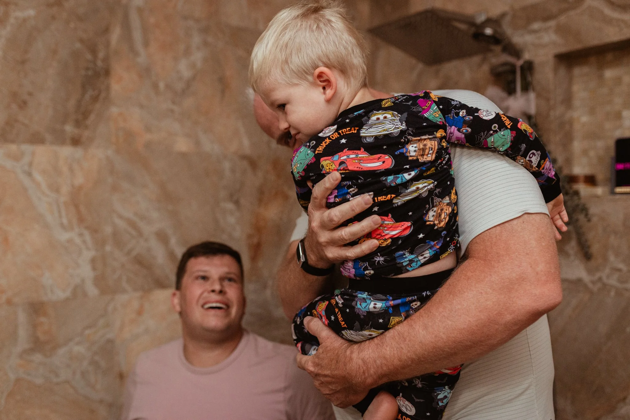A young boy in a robotics-themed shirt is lifted by an older person, while another person looks on and smiles in the background.