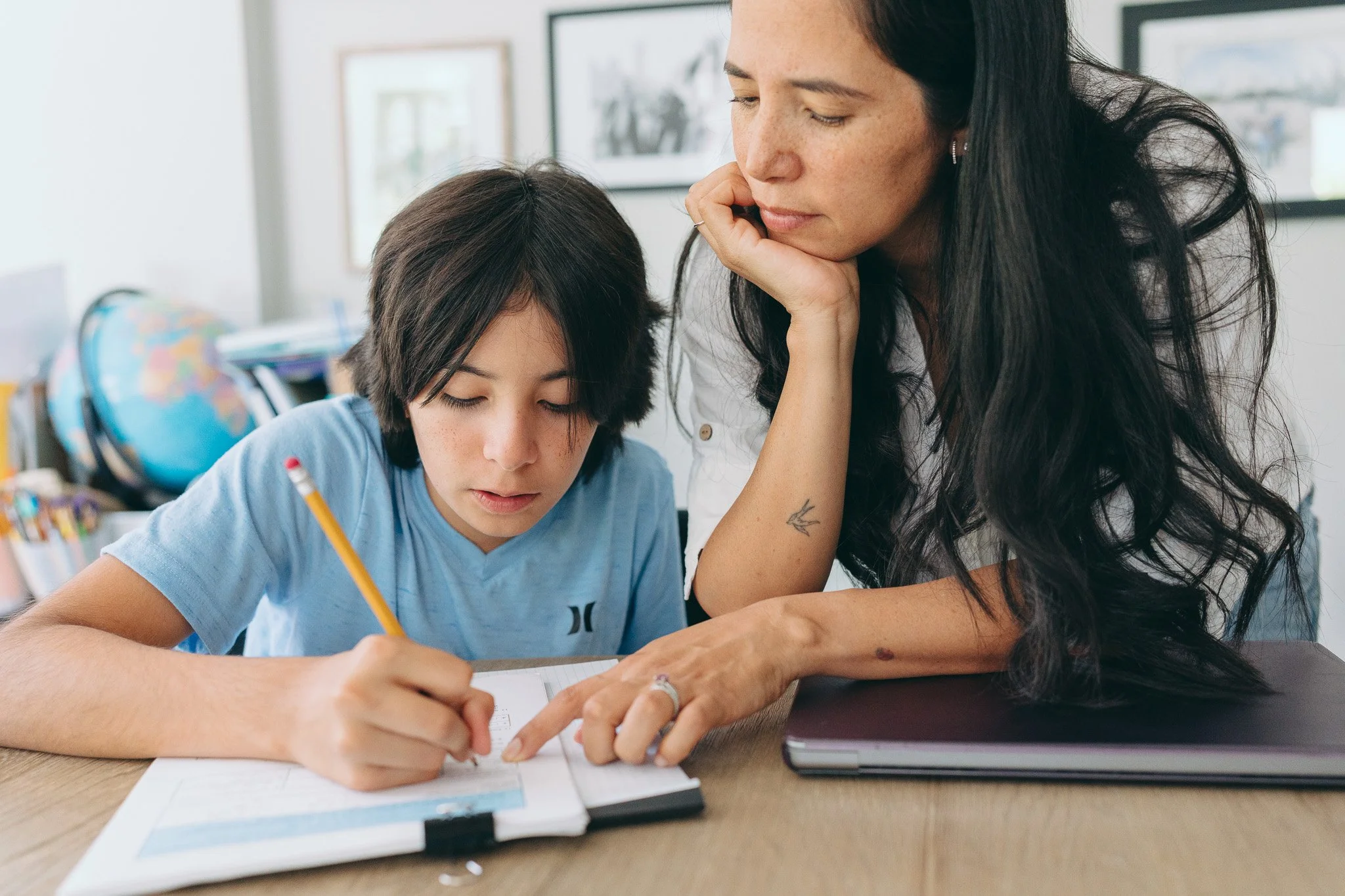 A woman with long dark hair and earrings assists a young boy with short dark hair in writing at a desk, with a globe and framed pictures in the background.