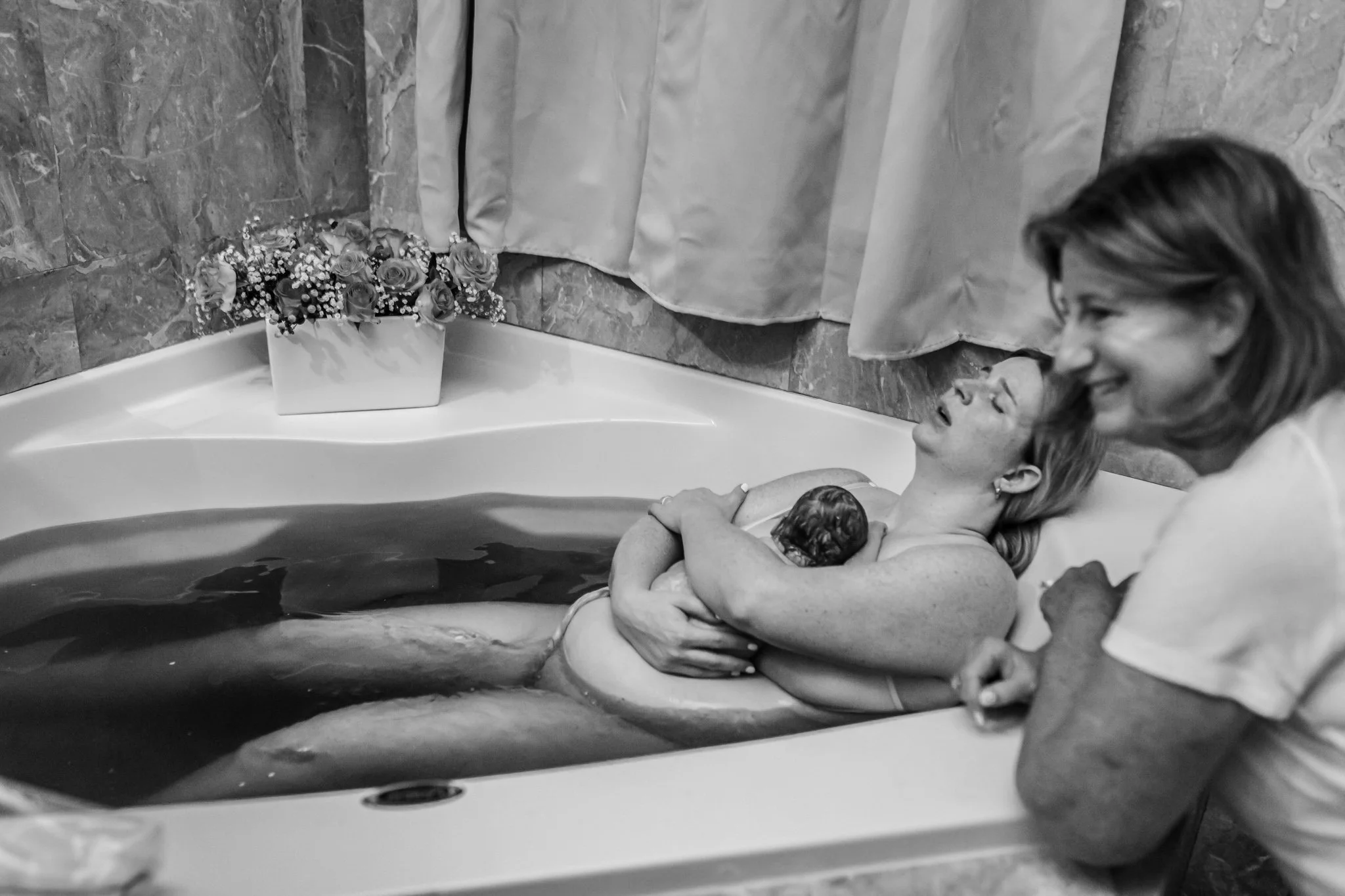 A woman holding a newborn baby in a bathtub with another woman leaning over her, all smiling, in a bathroom with a curtain and floral arrangement on the side.
