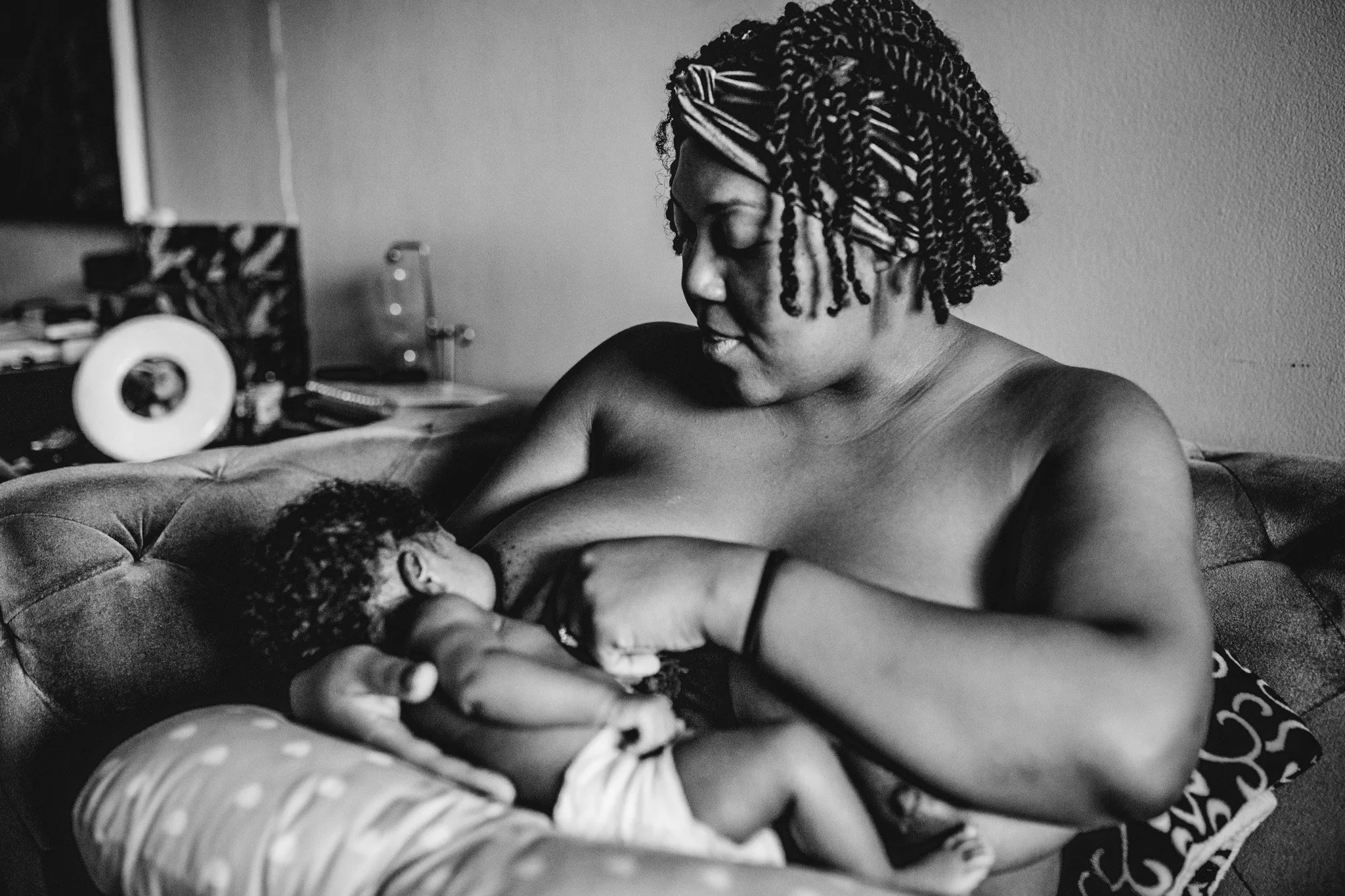 Black and white photo of a woman holding a baby, sitting on a couch. The woman has curly, styled hair with twists, and the baby is breastfeeding. The background includes a blurred wall and some household items.
