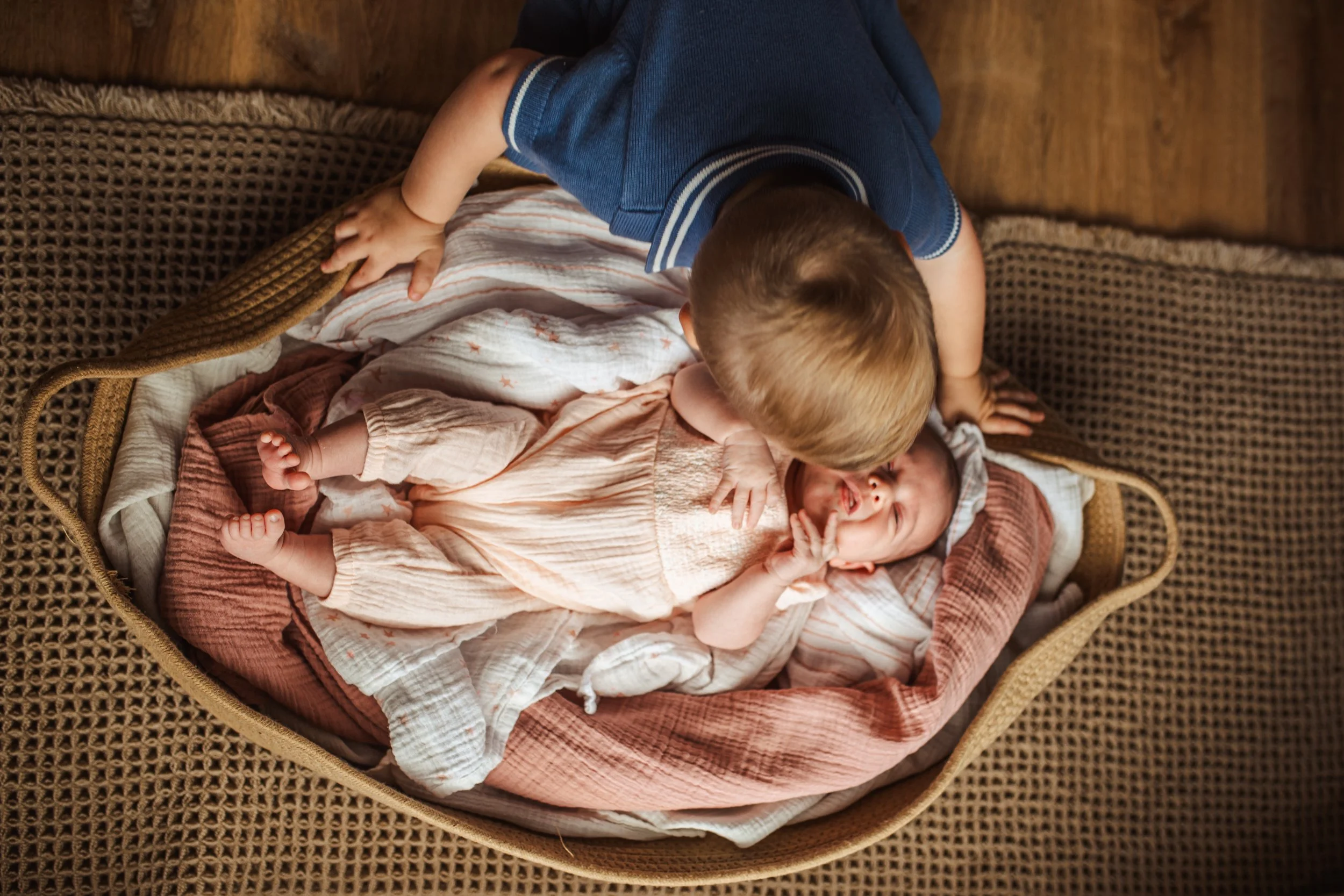 A young boy in a blue shirt is kissing or cuddling a laughing baby lying in a woven bassinet with a pink blanket, on a wooden floor.