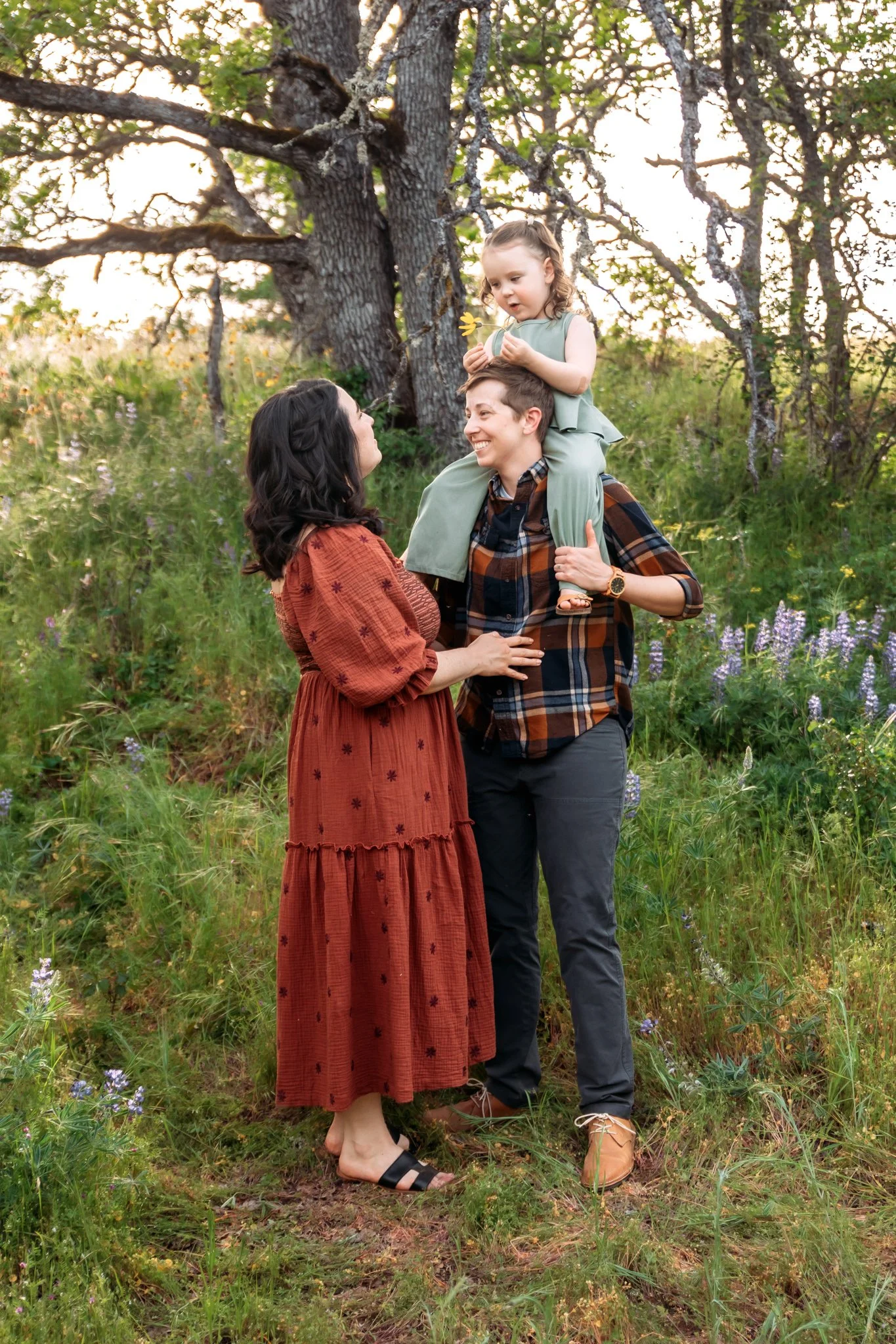 A family of three enjoying a walk in a forest clearing during sunset. The woman is wearing a rust-colored dress with embroidered details and sandals, the man is wearing a plaid shirt with dark pants and brown shoes, and the young girl, sitting on his