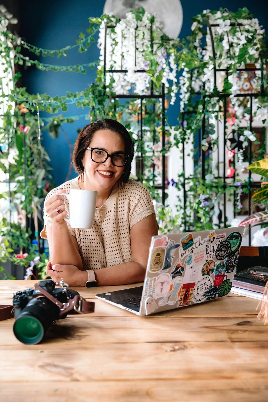 A woman with dark hair, glasses, and a beige crochet top sitting at a wooden table, holding a white mug, smiling at the camera, surrounded by plants and decorated with stickers on her laptop.