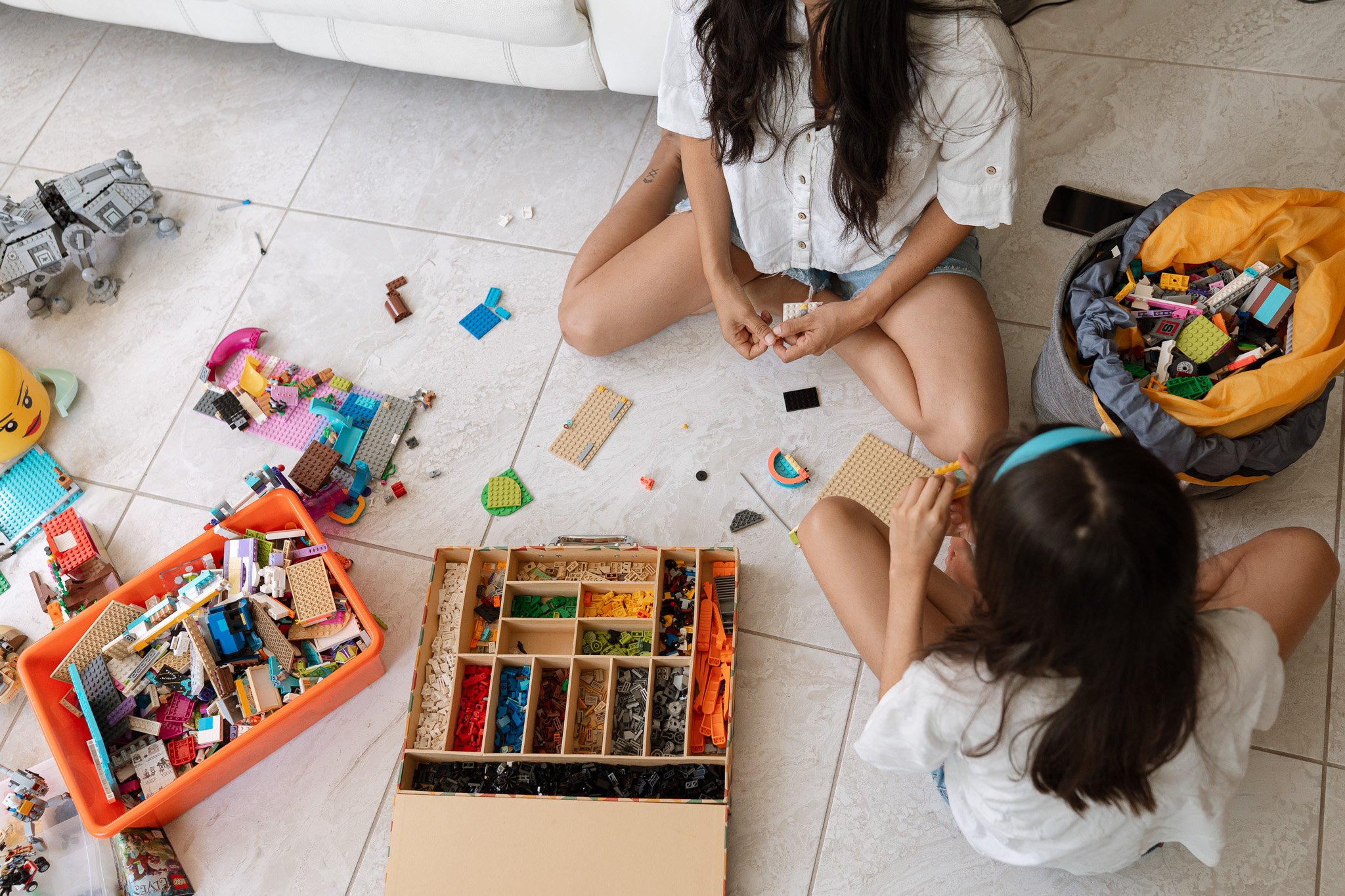 Two children sitting on a tiled floor playing with an assortment of LEGO bricks and building toys; a large LEGO set is in progress, and a bag filled with LEGO pieces is nearby.