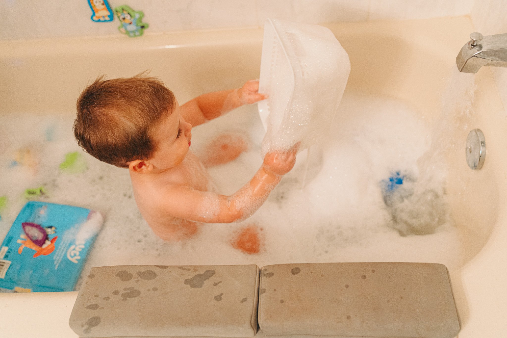A young child taking a bath in a bathtub, holding a large piece of bubble wrap. The tub is filled with soapy water and bubbles. There are cartoon character stickers on the wall and a box of bath toys nearby.