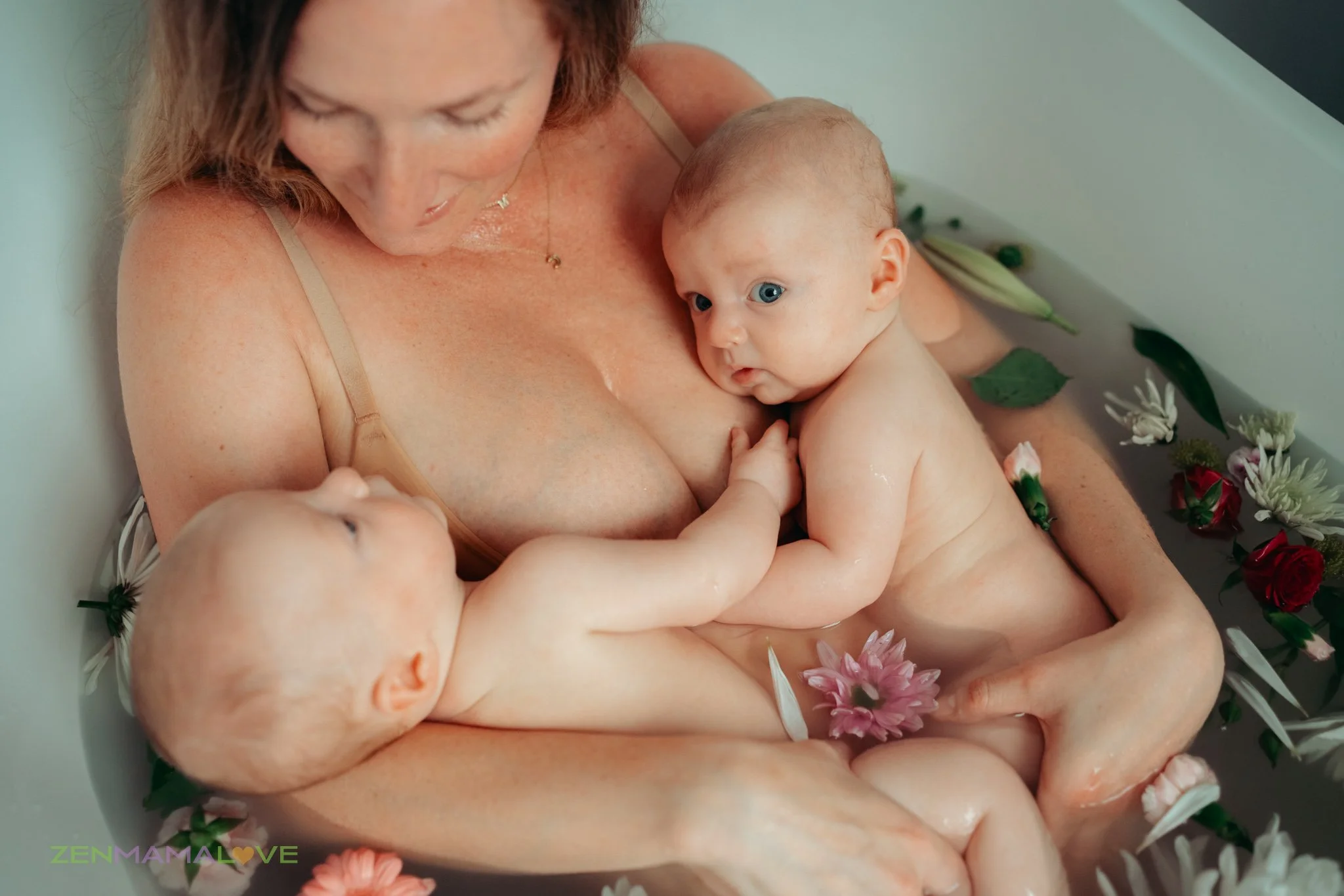 A woman holding two babies in a bathtub decorated with flowers, with one baby resting on her chest and the other boy looking at the camera.
