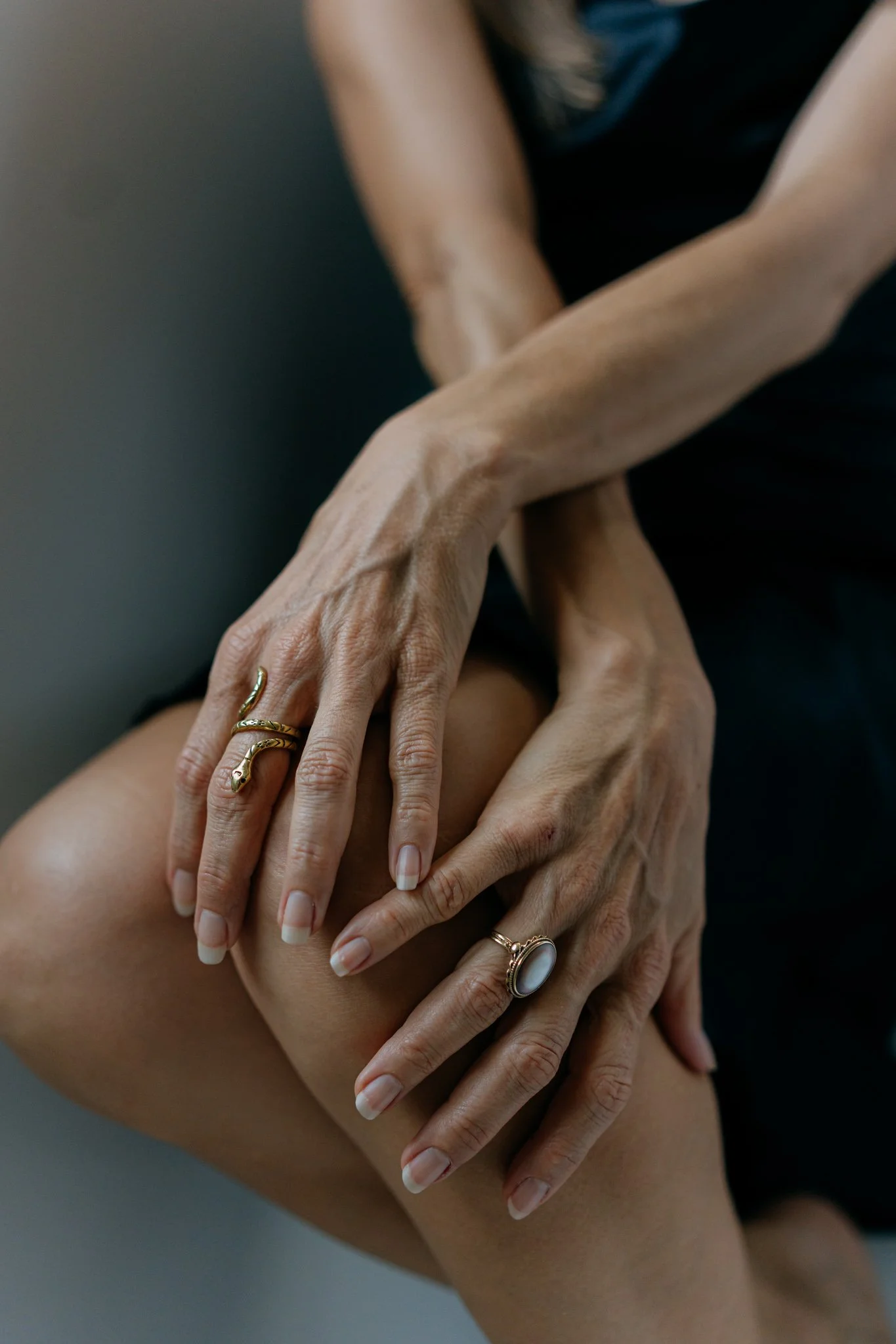 Close-up of hands with various rings, one with a snake design, gently holding another person's hand and arm. The background is blurred, emphasizing the hands.