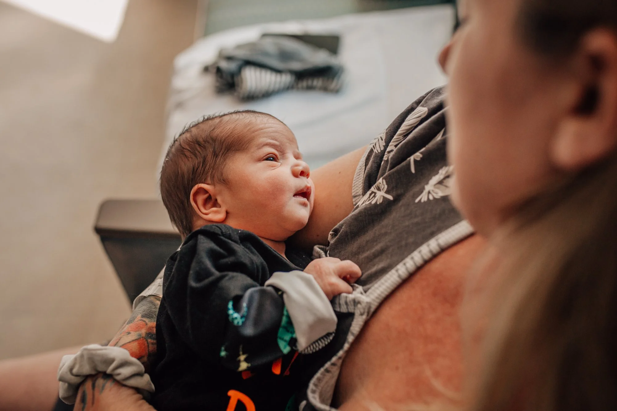 A close-up of a newborn baby with a woman, likely the mother, holding the baby on her chest. The baby is looking upward, and the woman's face is partially visible on the right side of the image.