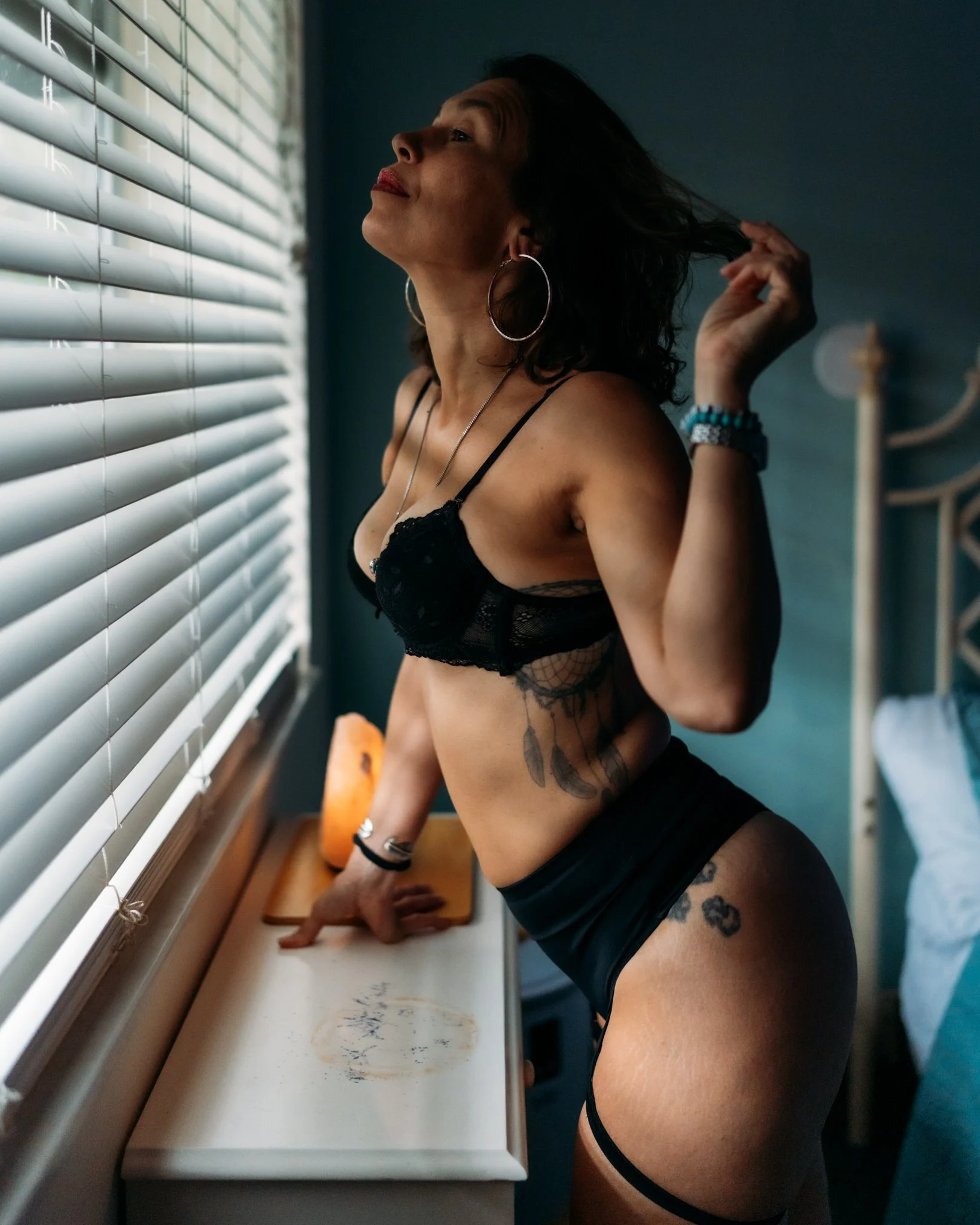 A woman with dark wavy hair, hoop earrings, and tattoos wearing black lace lingerie and black shorts, standing by a window with closed blinds, pulling her hair back while looking out the window in a dimly lit room.