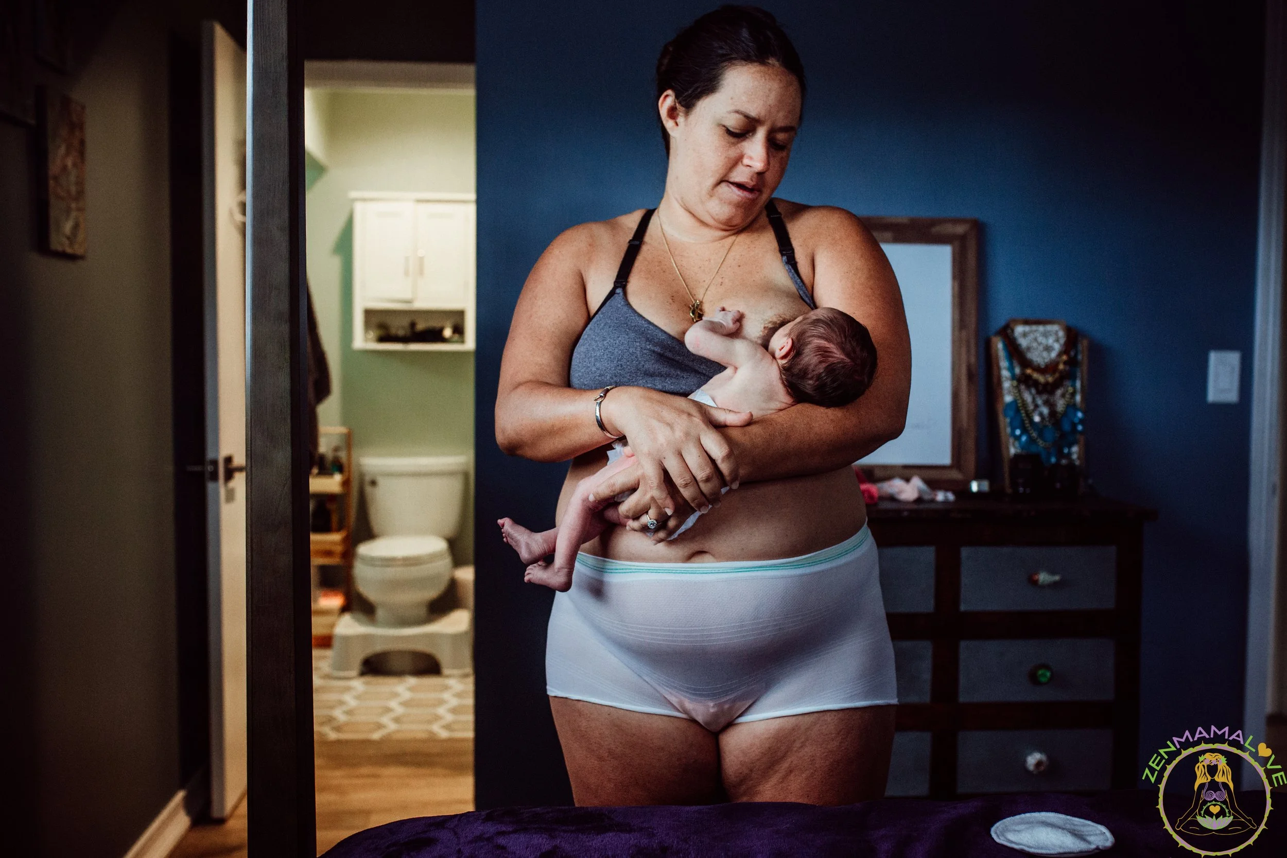 A woman wearing a tank top and underwear breastfeeding a baby in a room with blue walls, a mirror, and a decorated dresser in the background.