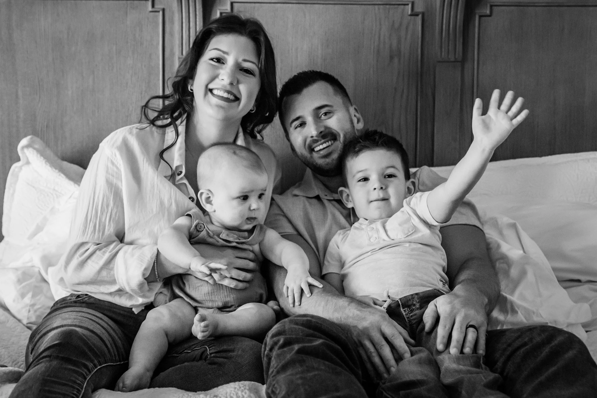 A black and white photo of a happy family sitting on a bed, waving at the camera. The group includes a woman, a man, and two young children, one of whom is sitting on the woman's lap and the other sitting on the man's lap. They are all smiling and ap