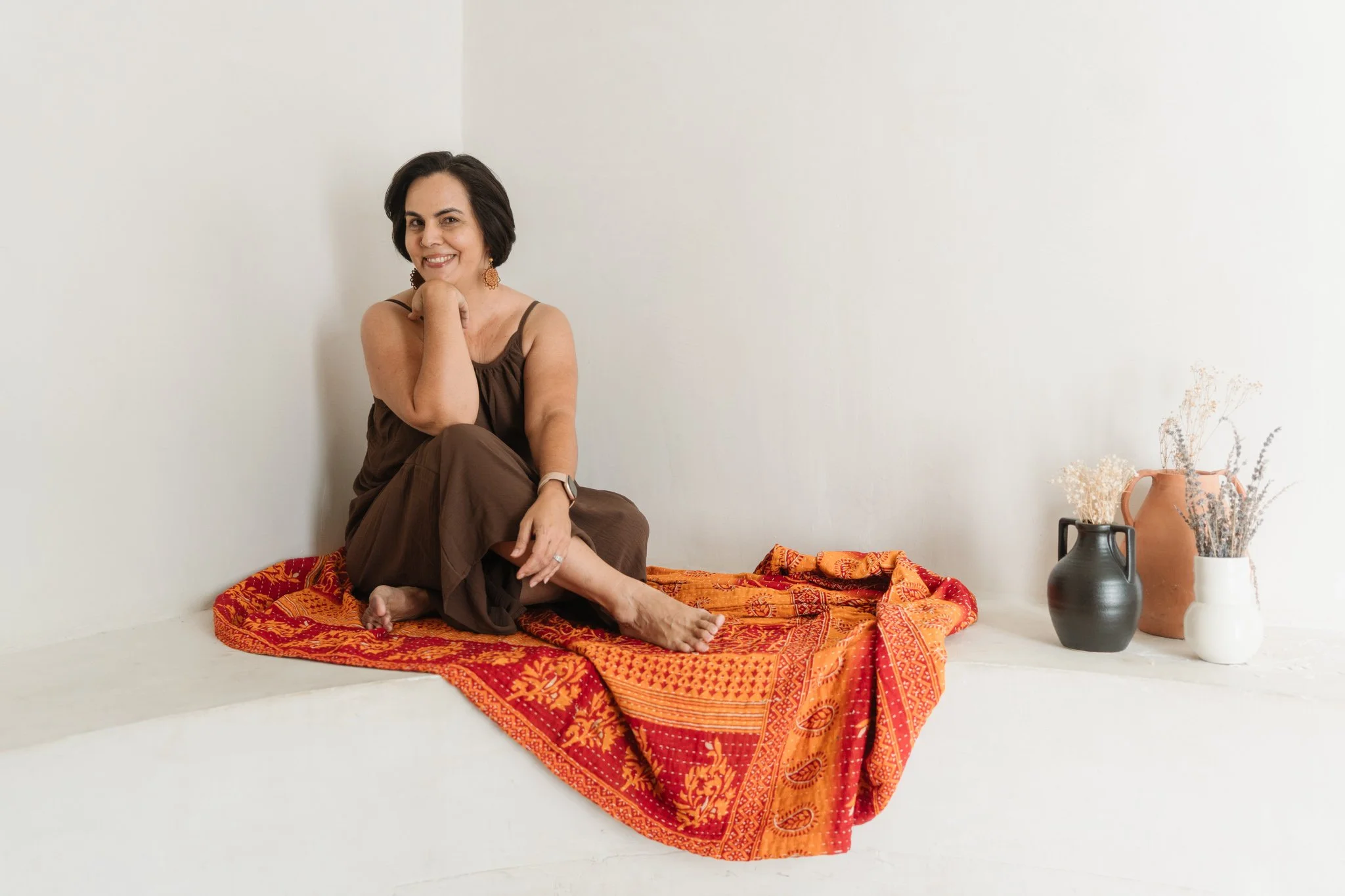 A woman with short dark hair, smiling, sitting on a red and orange patterned blanket on the floor, wearing a brown dress with spaghetti straps, in a minimalistic room with white walls. There are various vases with dried flowers placed nearby.