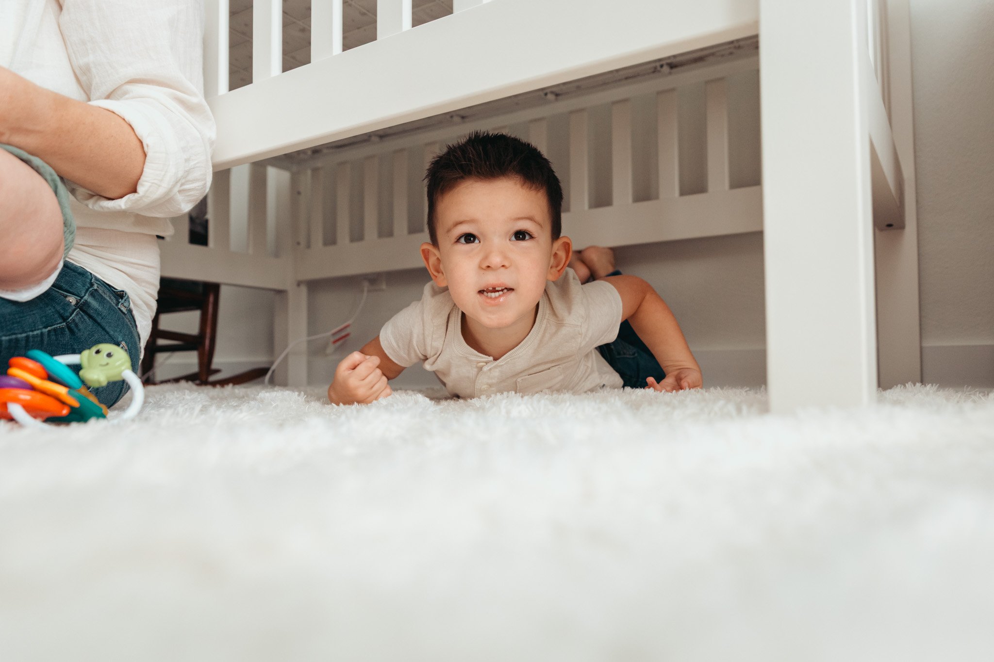 Young boy crawling under a white bed with a white rug and a colorful toy on the floor.