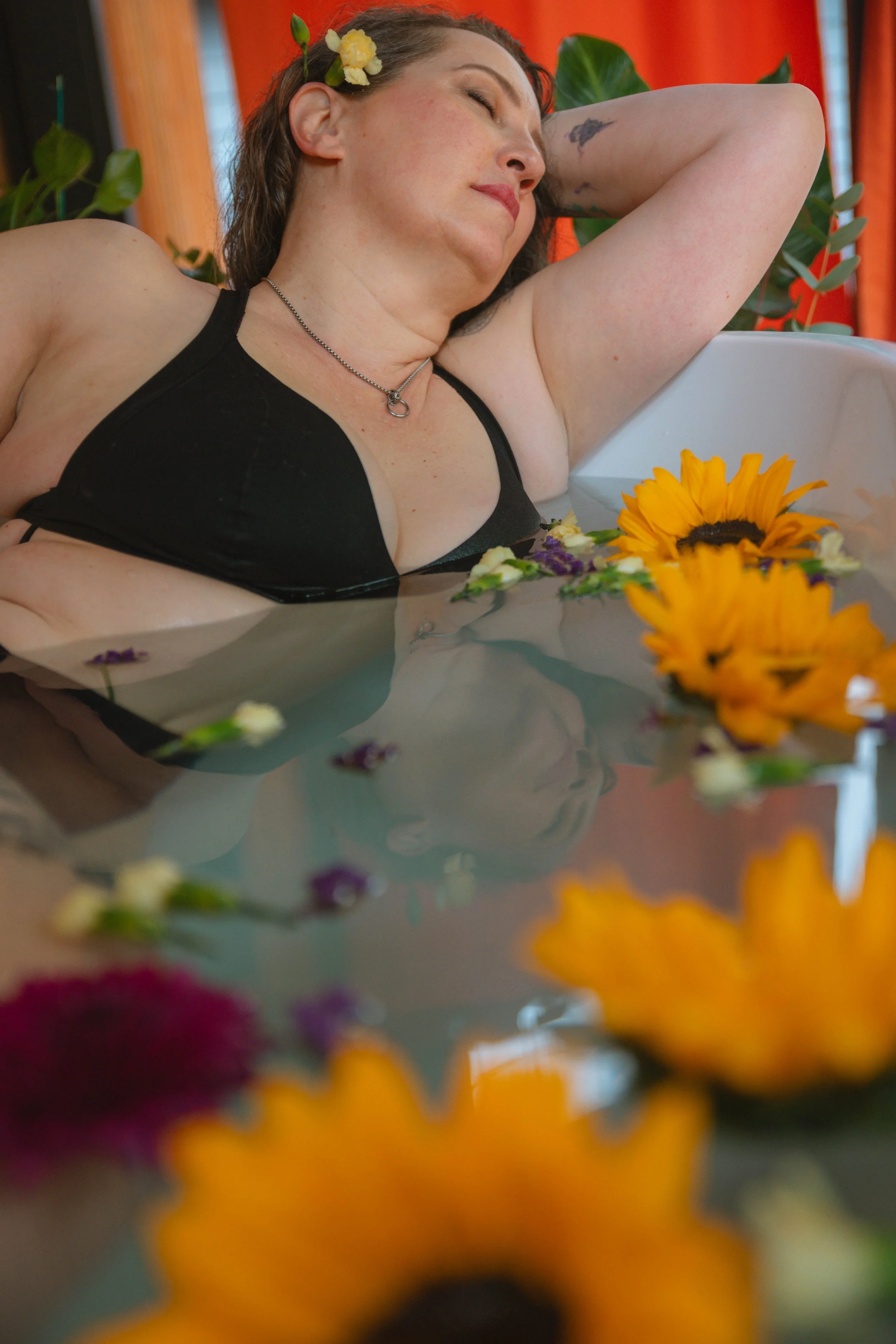 A woman with dark hair, a flower in her hair, relaxing with her eyes closed in a bathtub surrounded by colorful flowers and green plants.