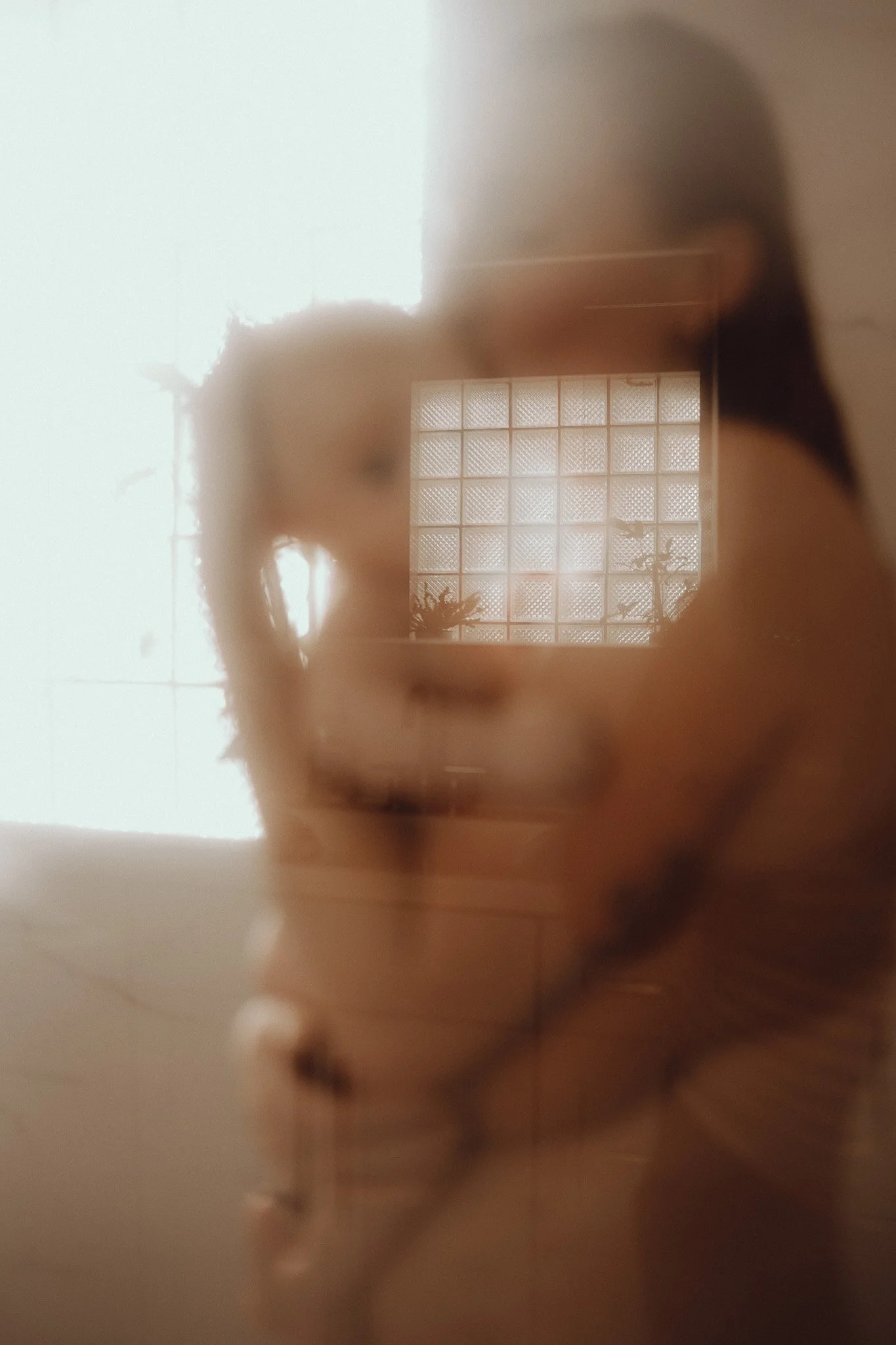 A person taking a mirror selfie in a bathroom with frosted glass block windows, showing their silhouette and hair.