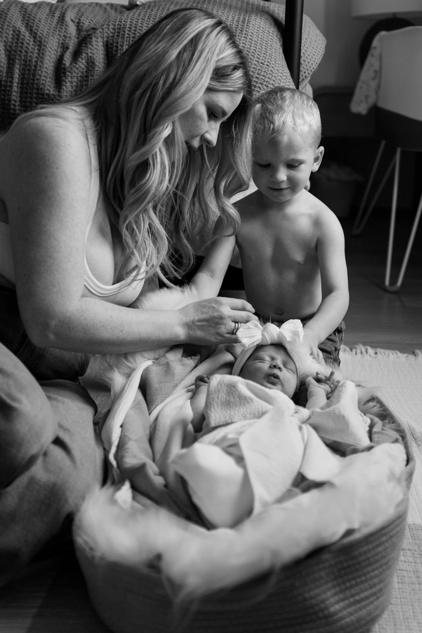 A woman with long, wavy hair and light skin sits on the floor with two young children, one standing and one lying in a blanket. The woman gently touches the child's head, and they all look down at the baby, creating an intimate family moment in a coz